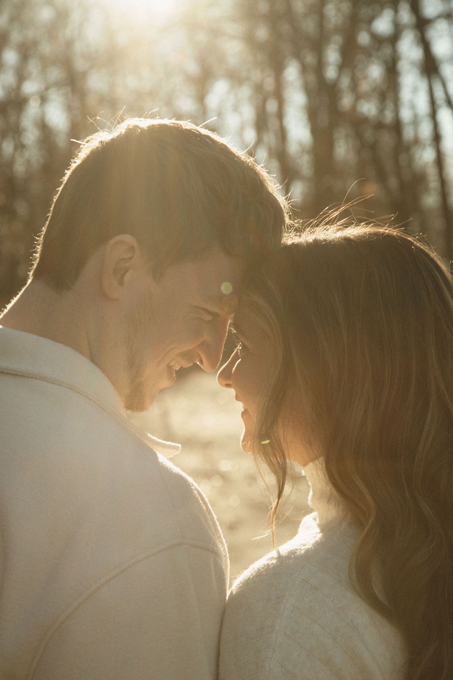 A backlit couple sharing a tender moment at sunset in a wooded area.