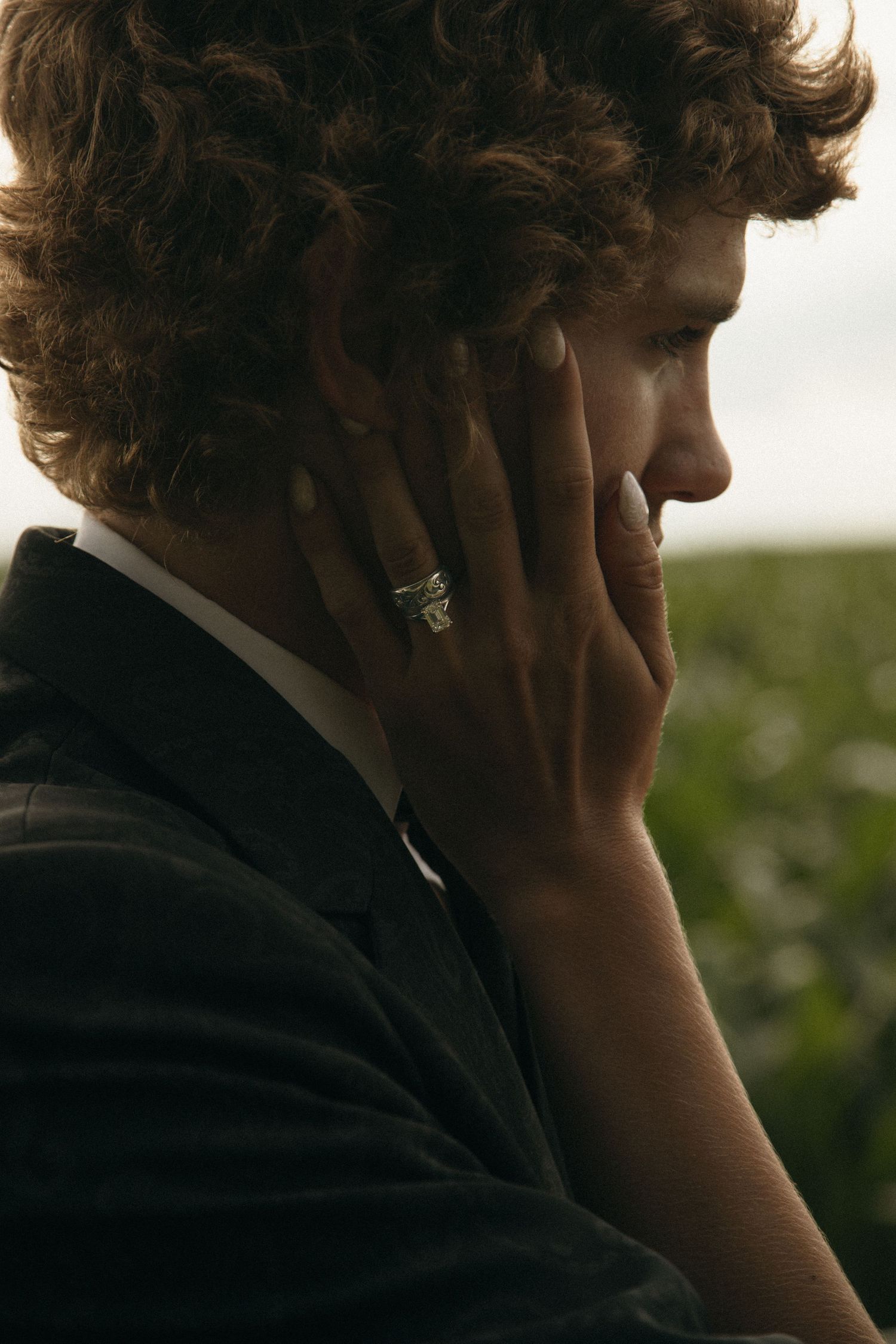 Close-up side profile of curly-haired groom in dark suit with bride's hand showing wedding ring.