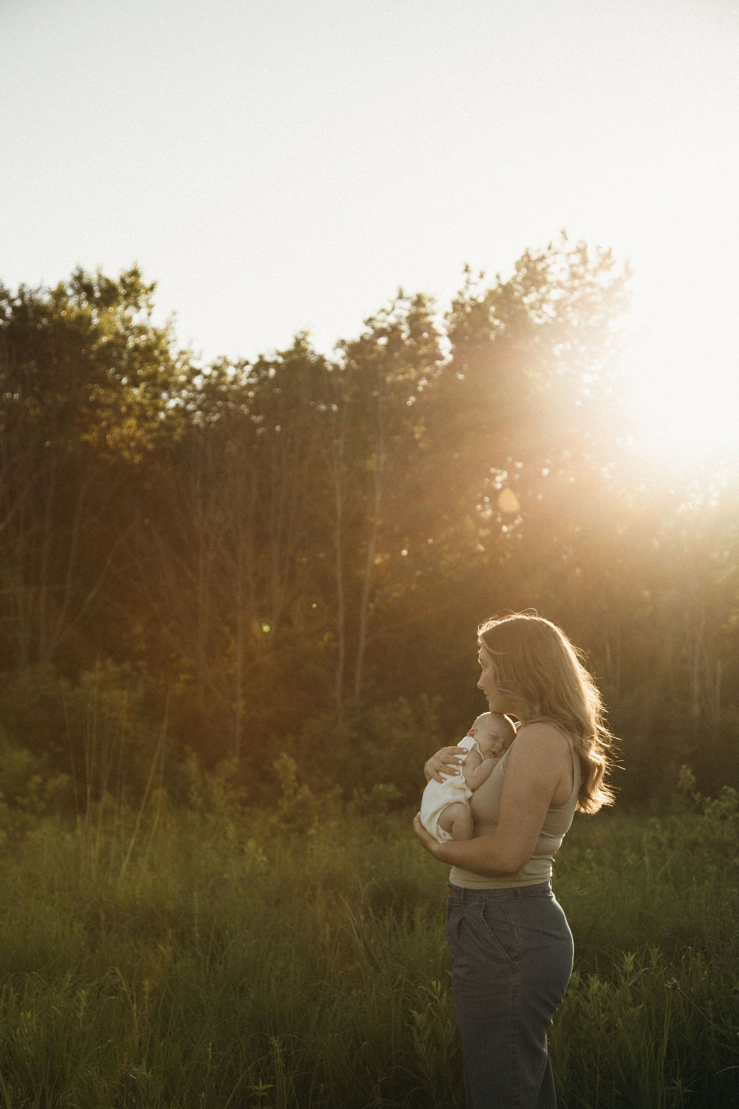 Mother cradling newborn baby in a sunlit green field with glowing backlight.