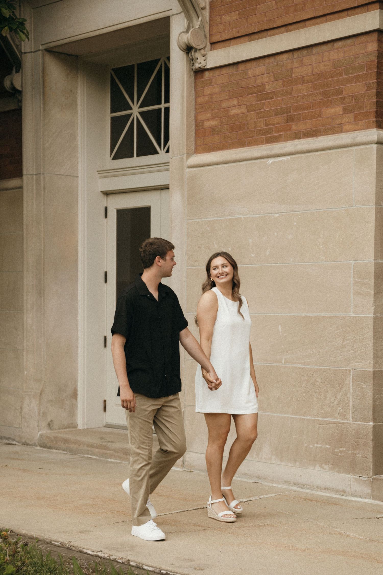 A smiling couple holds hands while walking outside a brick building, woman in white dress, man in black shirt.