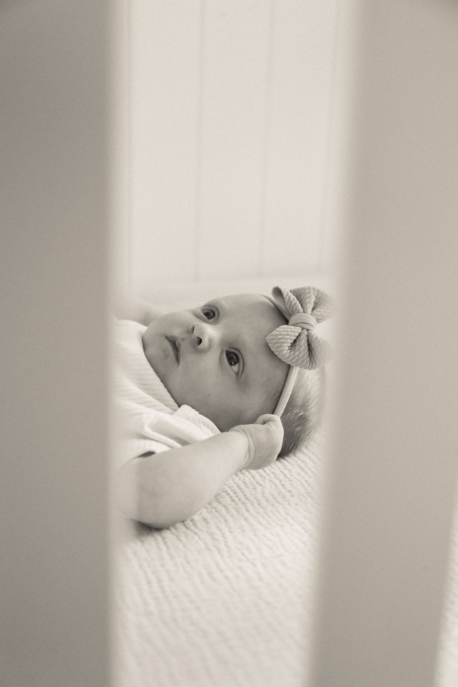A baby lies in a white crib viewed through the slats in black and white photography.