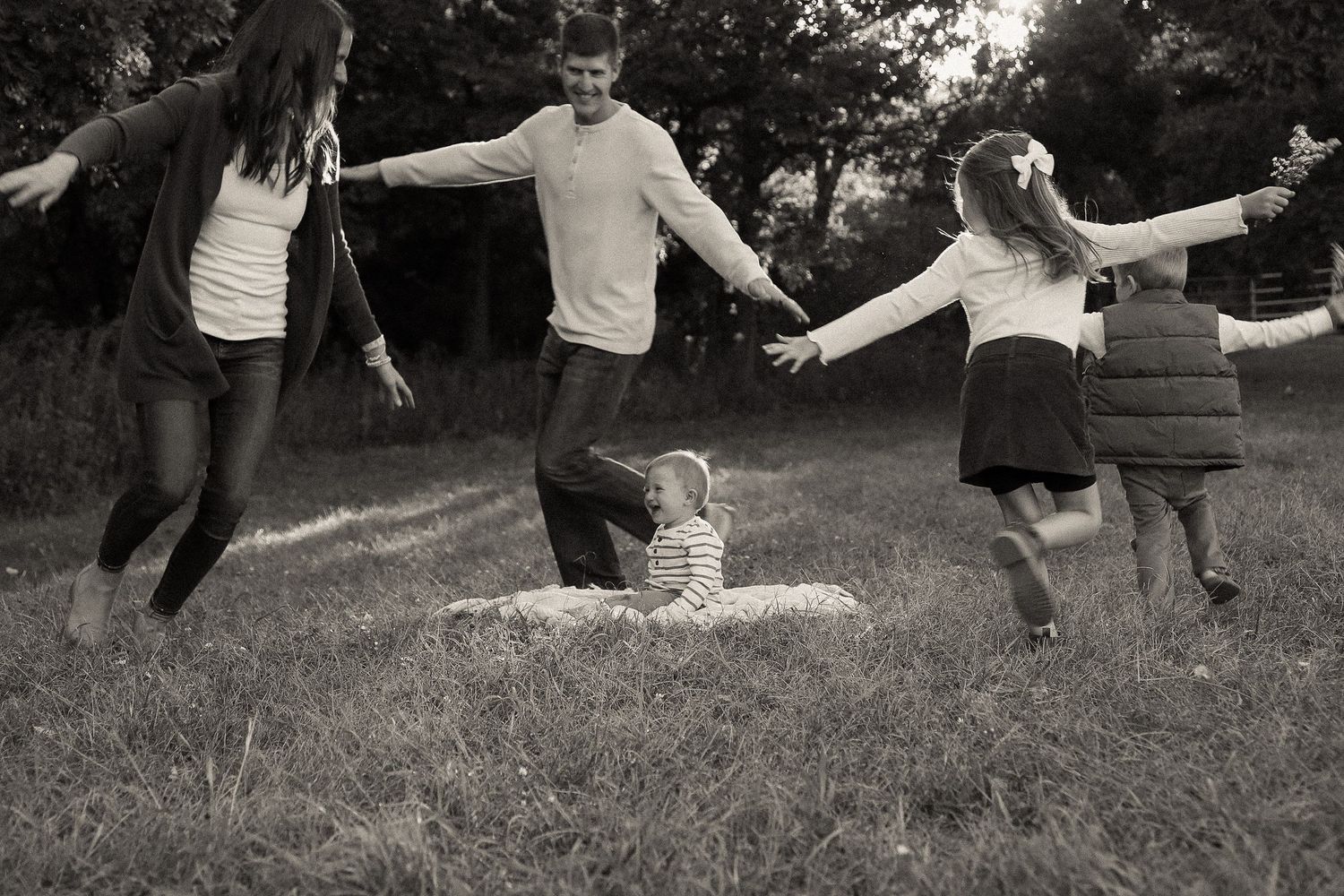 Family plays together on a grassy hillside during golden hour, creating a playful silhouette against the sunset.