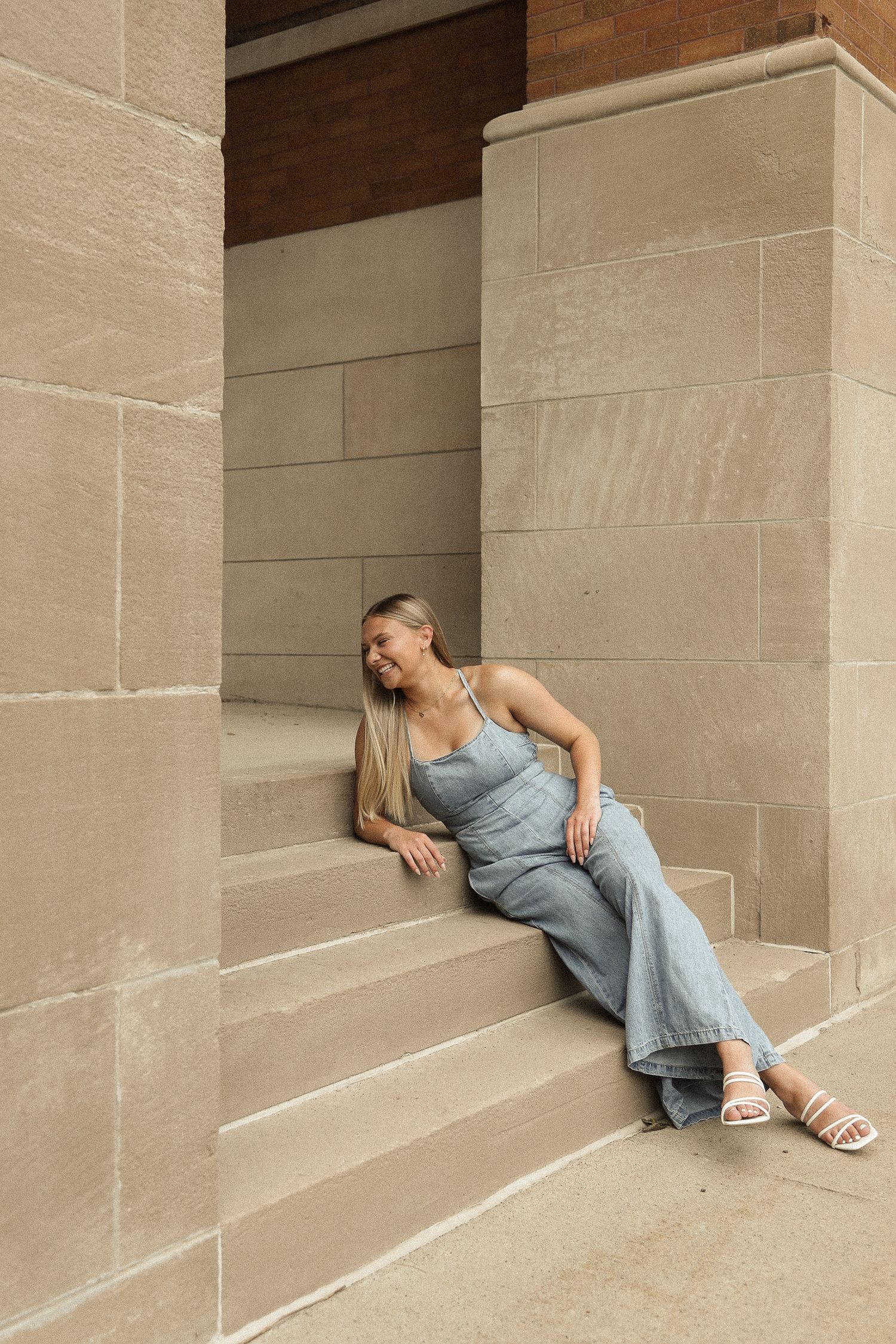 Figure lounging against stone wall in casual denim outfit and sandals in architectural setting.