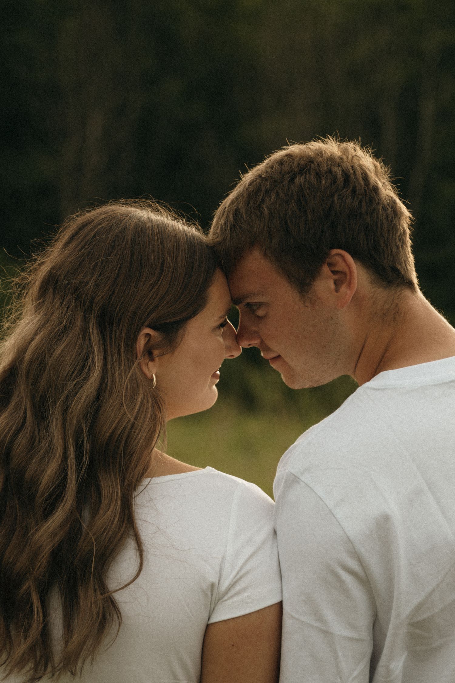 A smiling couple in white t-shirts touch foreheads intimately outdoors with lush green trees in the background.