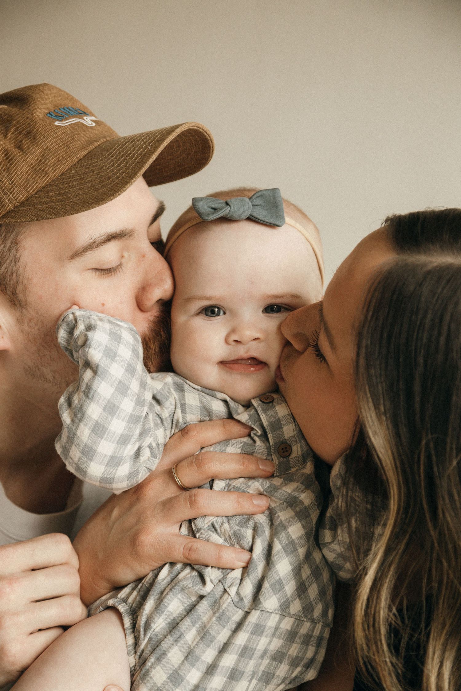 Two parents lovingly kiss their smiling baby wearing a gingham outfit and blue bow headband.