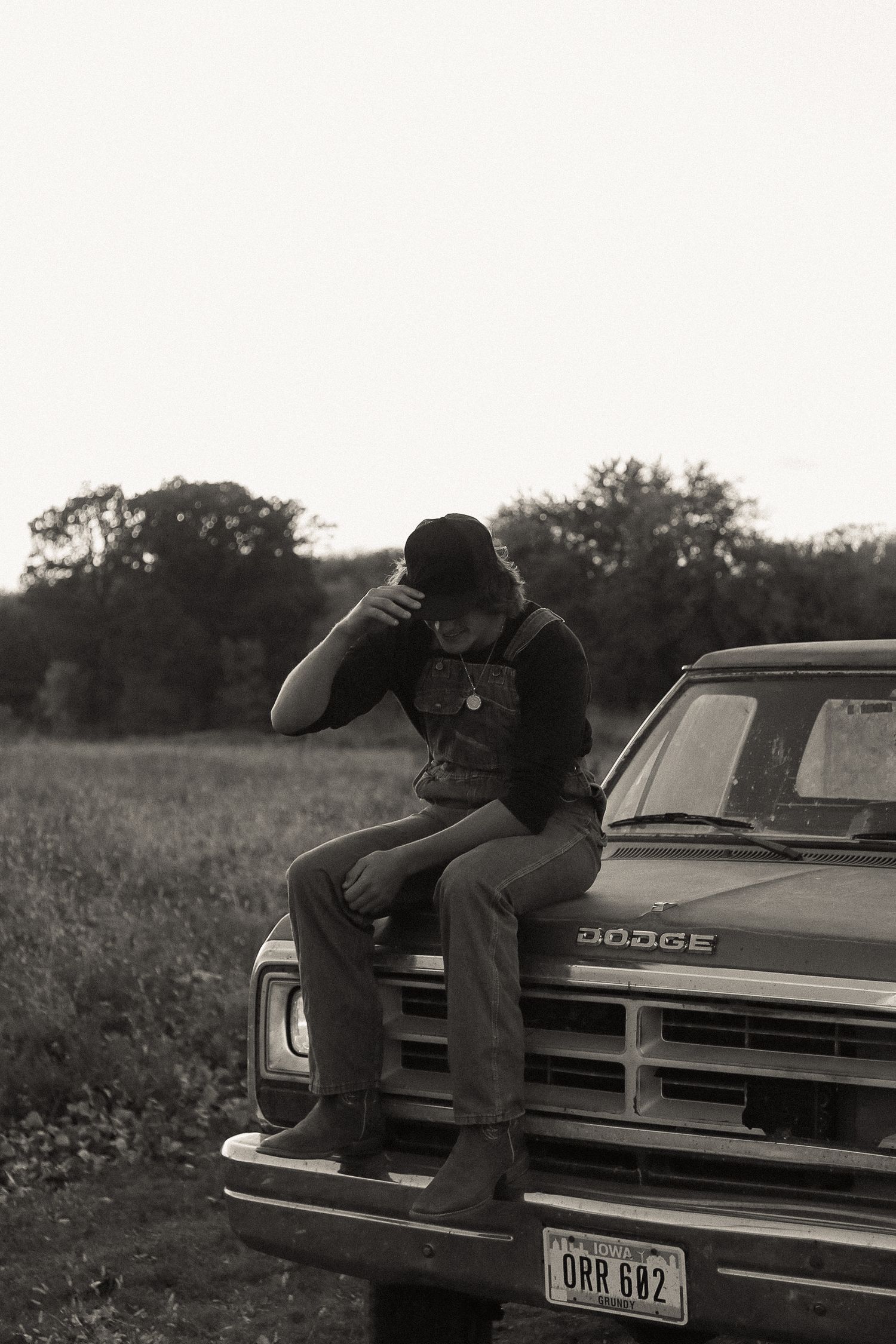 Figure sits on hood of classic Dodge car with license plate visible in rural setting.