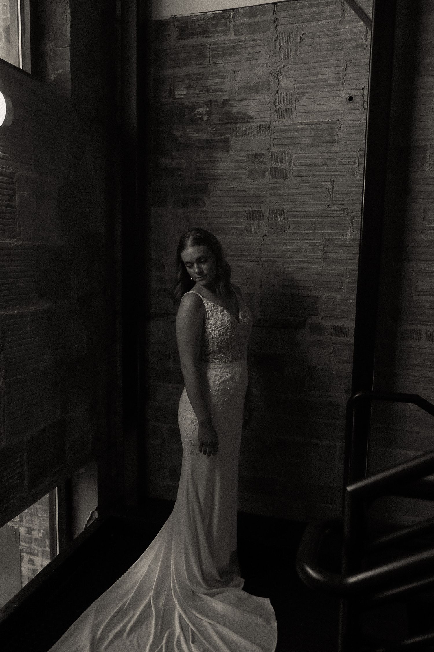 Person in a white lace dress standing against a brick wall in dramatic lighting.