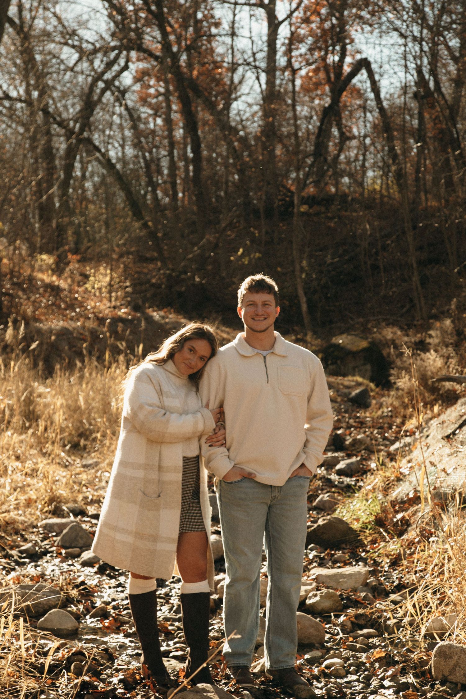 A cozy autumn portrait of a couple standing among fallen leaves and rocks in a wooded setting.