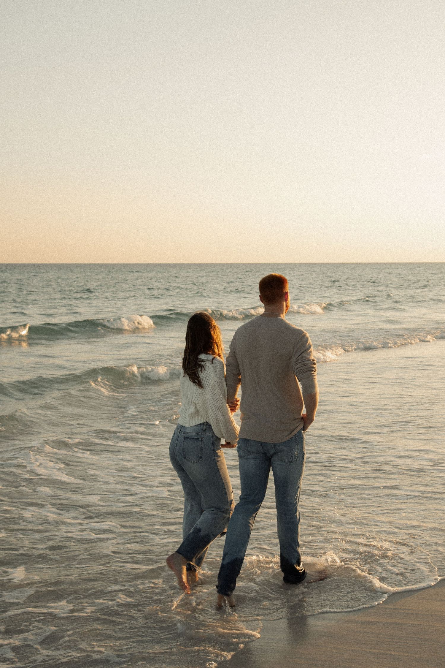 Couple walking together in shallow ocean waves at dusk.