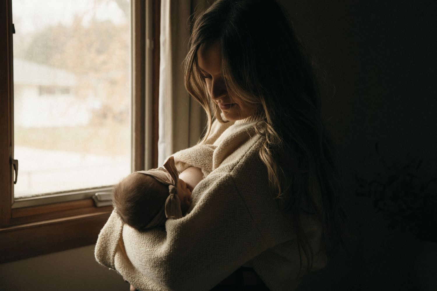 Silhouette of mother cradling newborn baby by window in moody lighting.