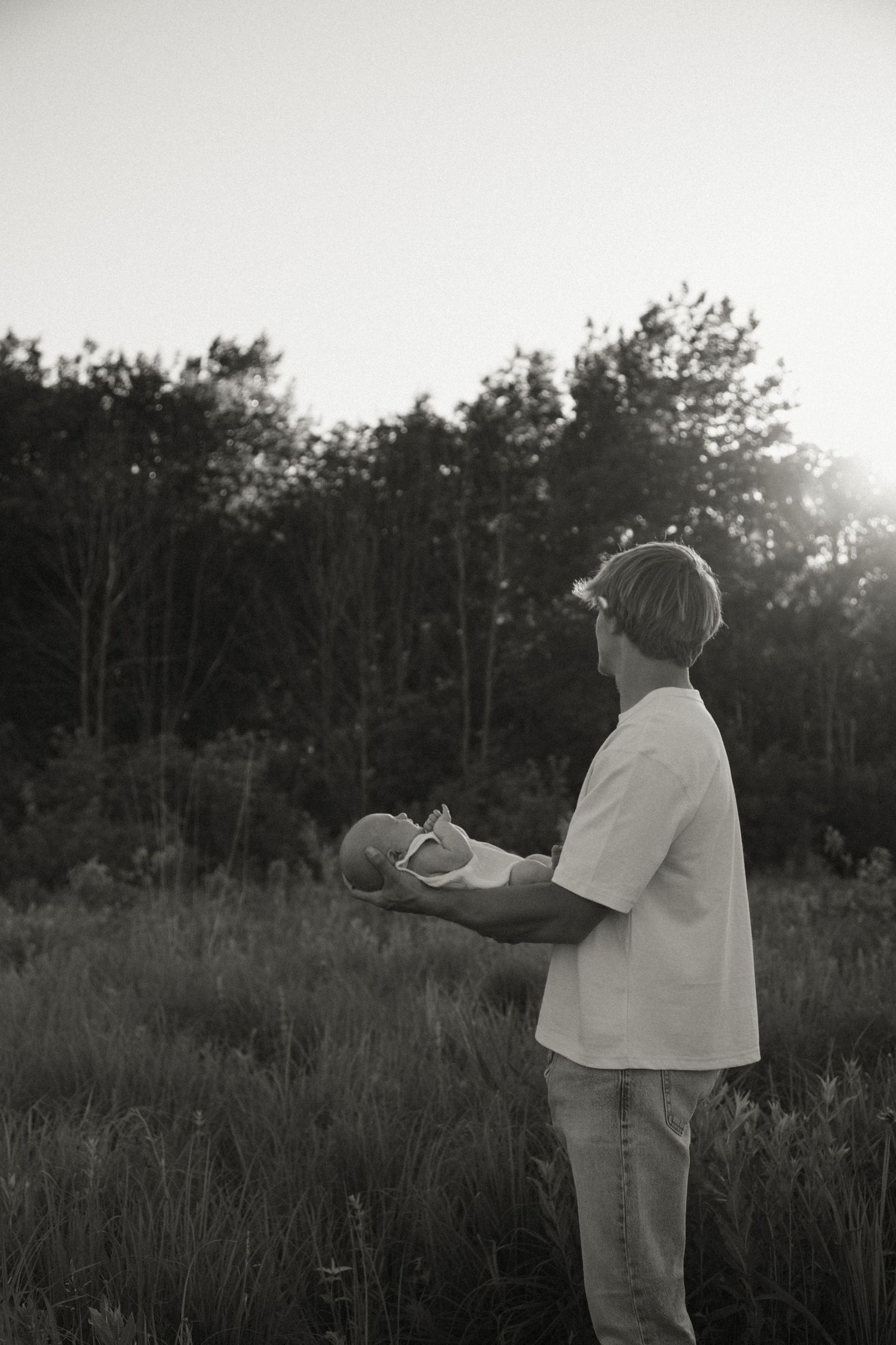 A father holds his newborn baby in a sunlit field with trees in the background, captured in black and white.