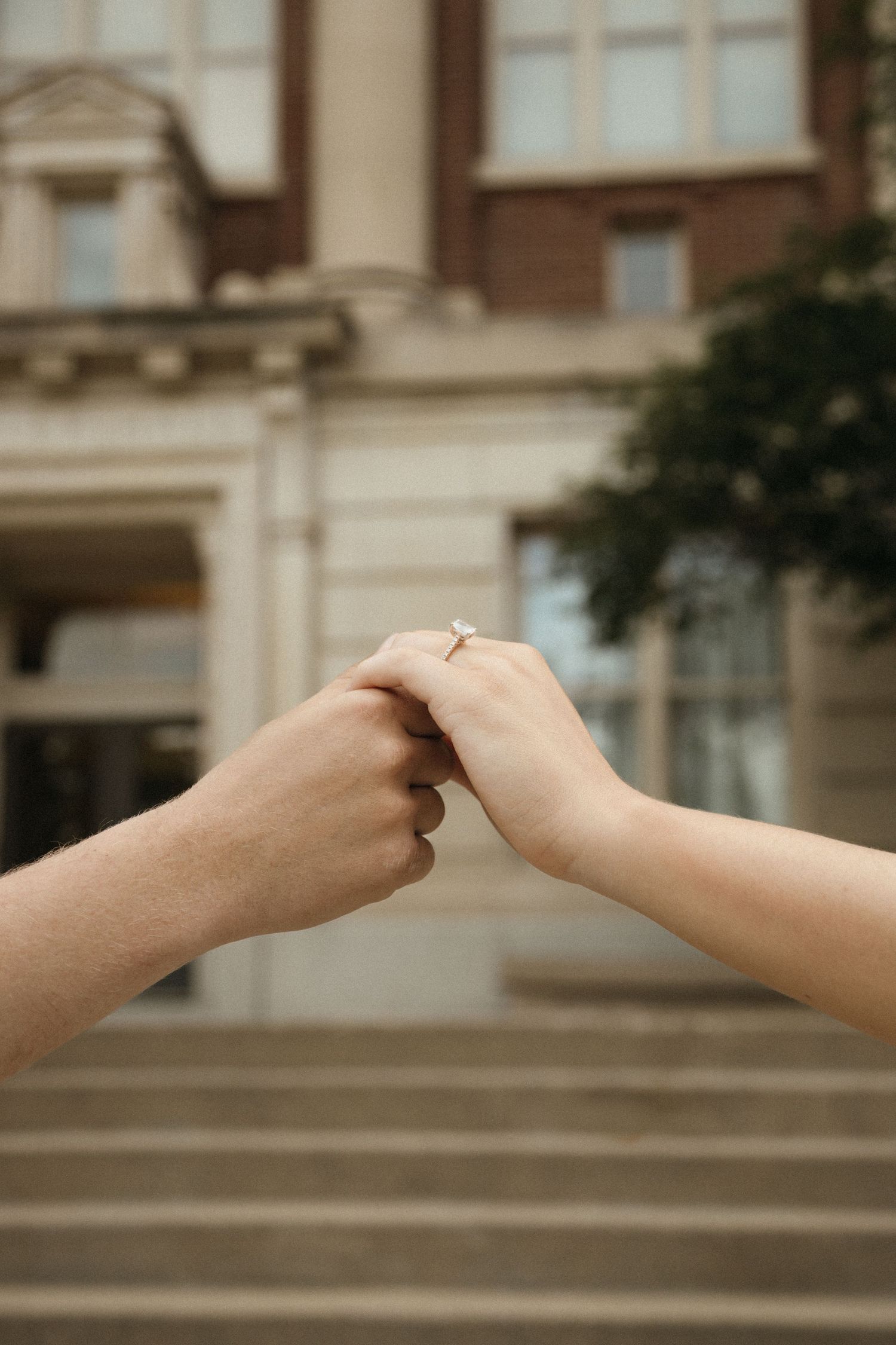 Close-up of two hands intertwined showing an engagement ring in front of a blurred brick building.