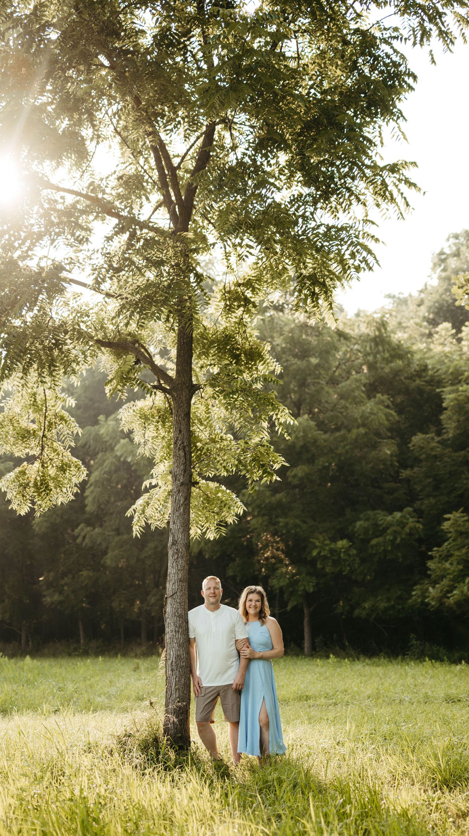 Couple standing together under a tall tree in a sunlit green meadow, woman in blue dress, golden hour lighting.