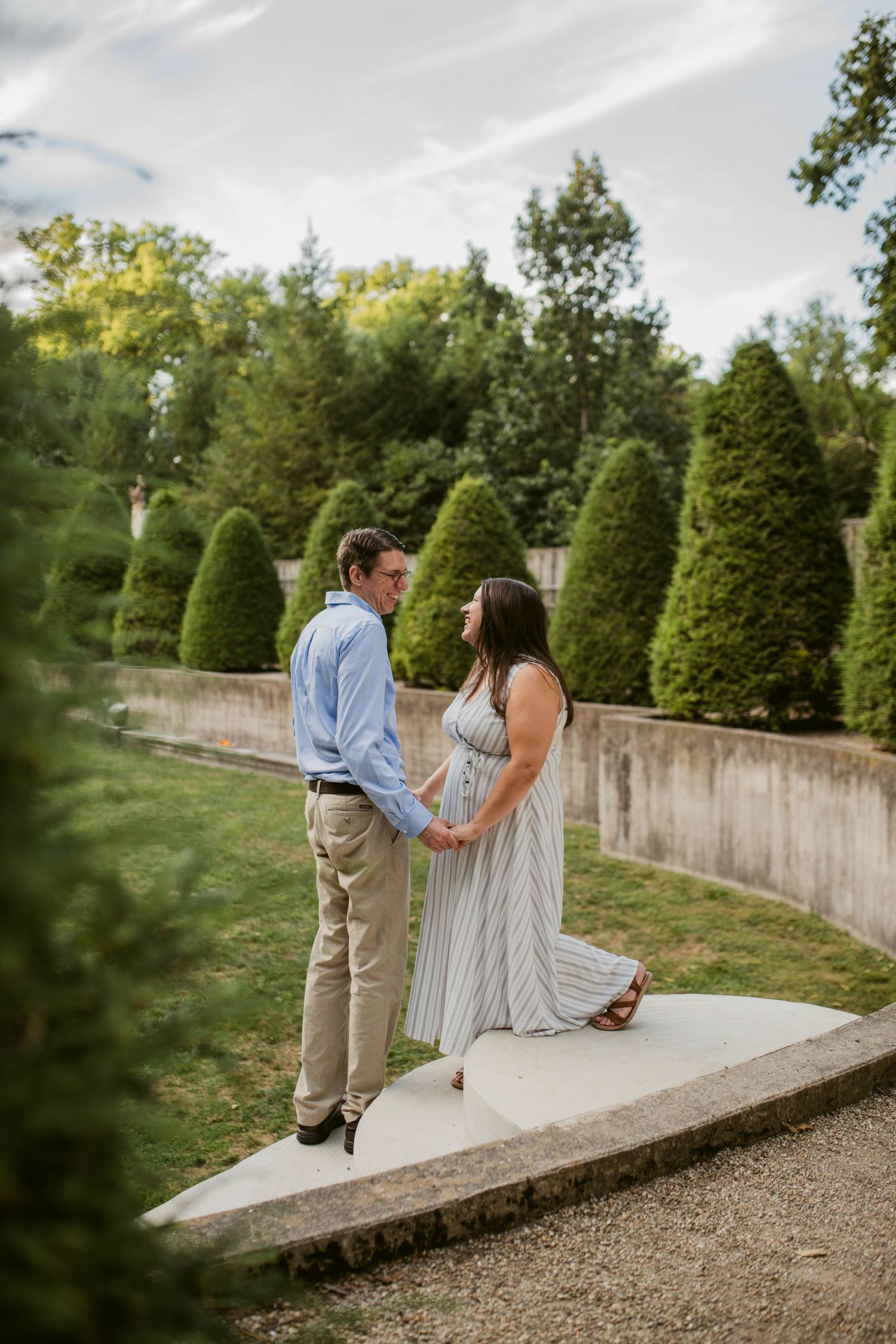 Couple holds hands facing each other on a garden path surrounded by manicured hedges and lush greenery.
central Illinois