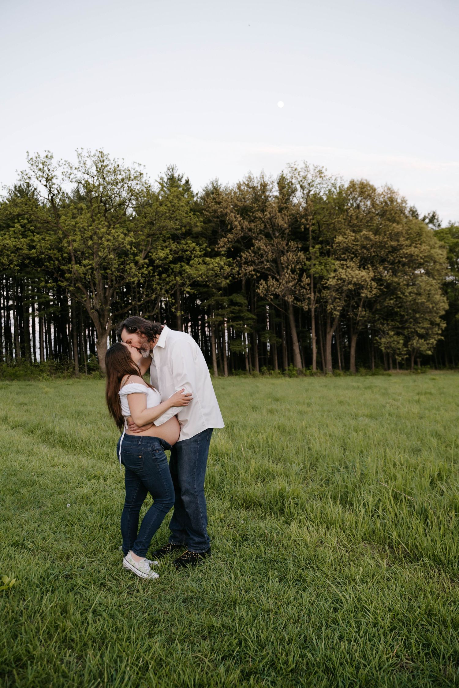 Couple sharing a romantic kiss in a lush green field with tall trees in the background during golden hour.