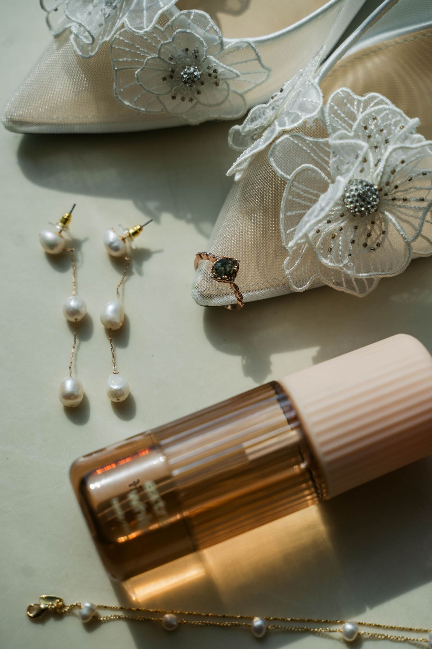 Bridal flat lay with pearl drop earrings, floral lace heels, gold necklace, and a peach beauty serum bottle.
central Illinois