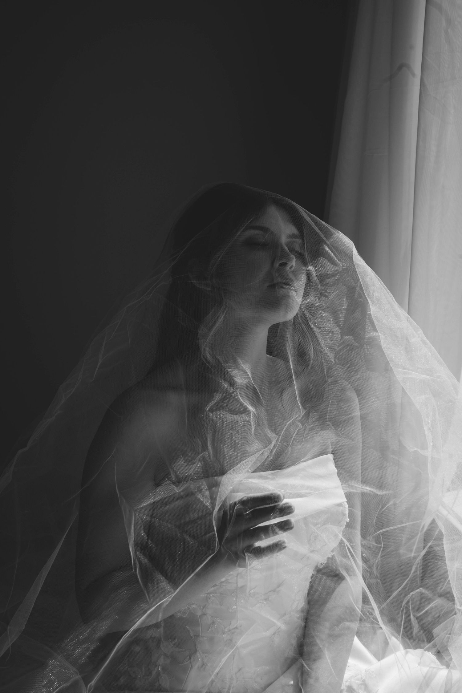 Black and white portrait of a bride in a wedding dress and veil by a window, gazing upward in soft natural light. central Illinois