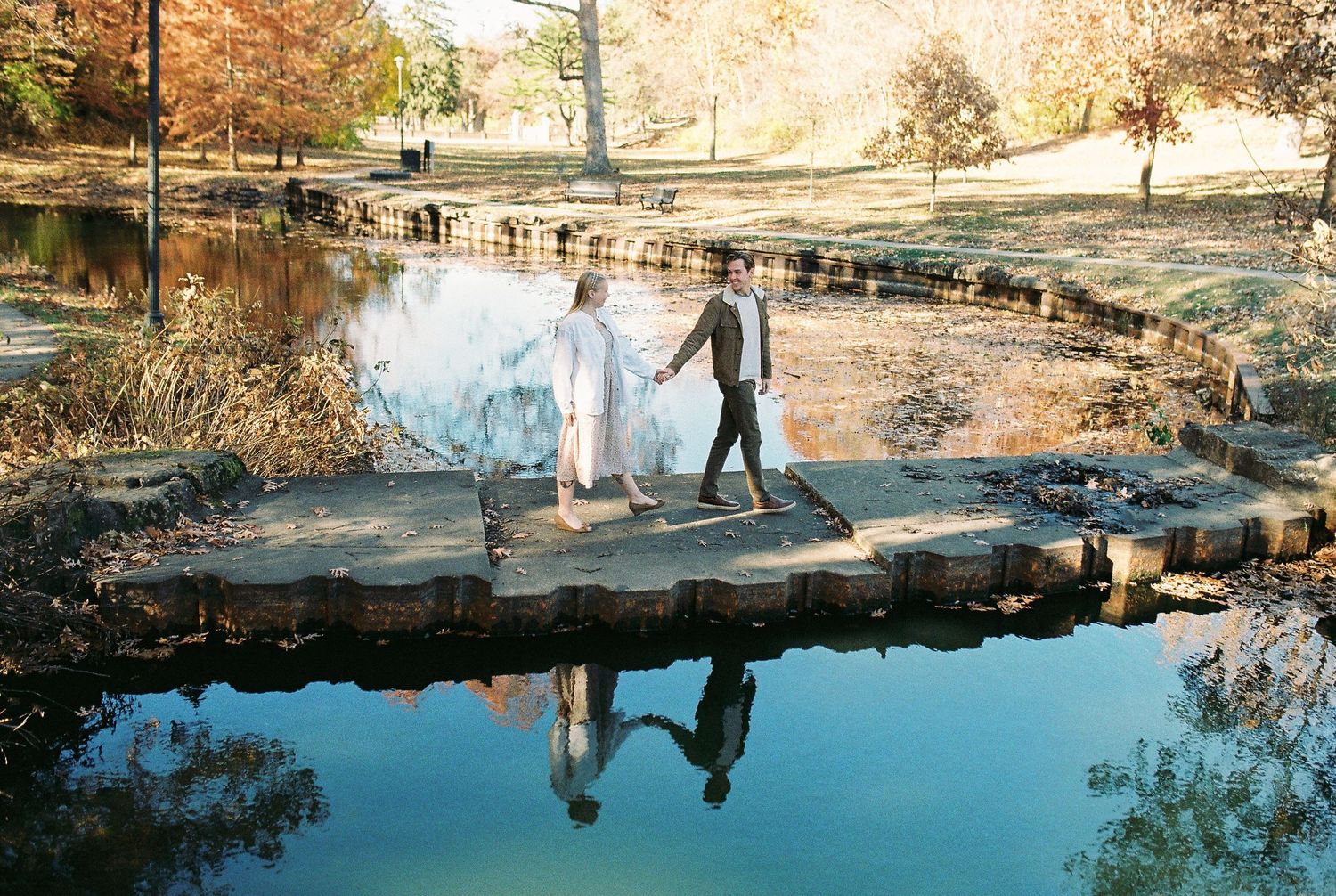 Couple holding hands walking on stone bridge over reflective pond in autumn park with colorful foliage.