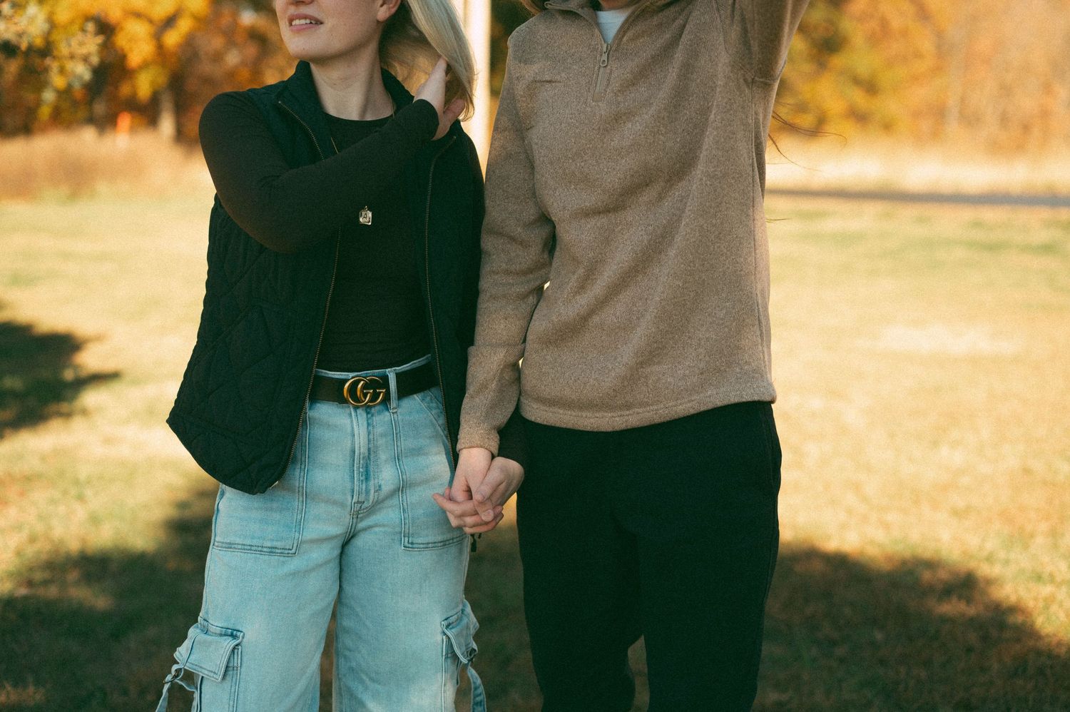 Couple holding hands outdoors in autumn park, woman in black blazer and jeans, man in beige sweater.