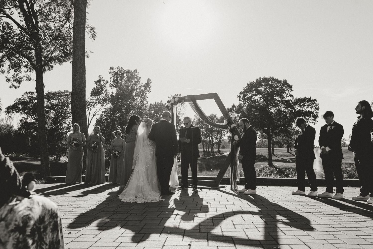 Black and white outdoor wedding ceremony with bride and groom under arch, guests standing on brick patio surrounded by trees.
illinois