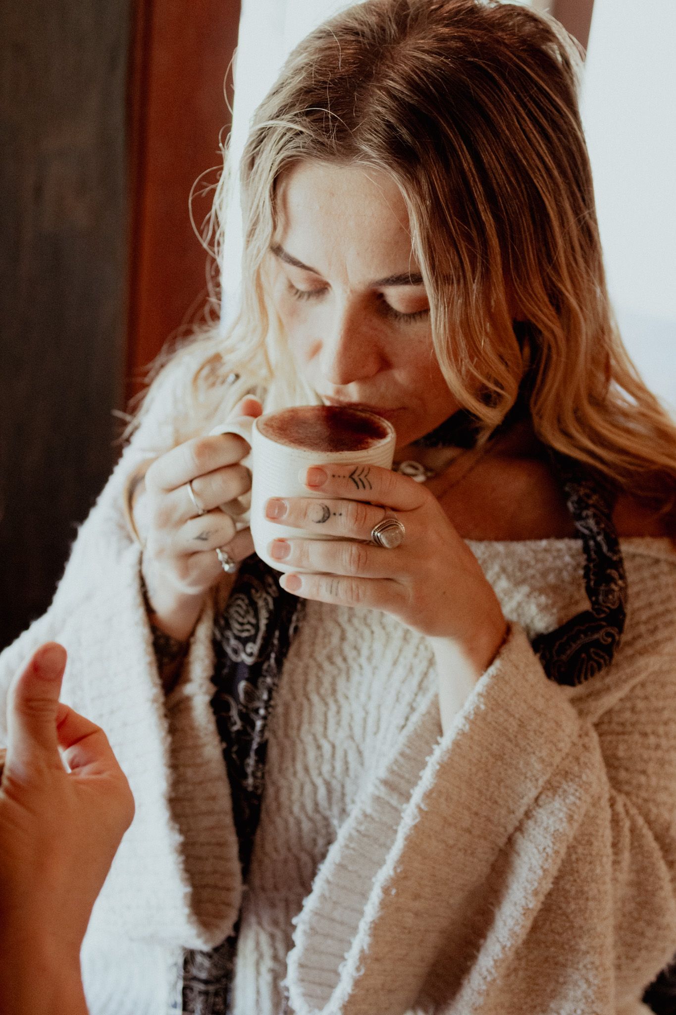 Person in cream knit sweater savoring a hot drink near a window.