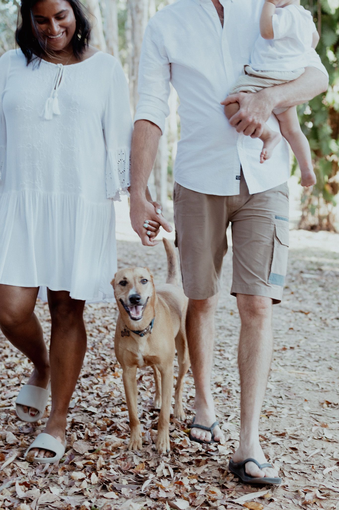 A family and their dog pose together outdoors wearing coordinated white outfits on a leaf-covered path.