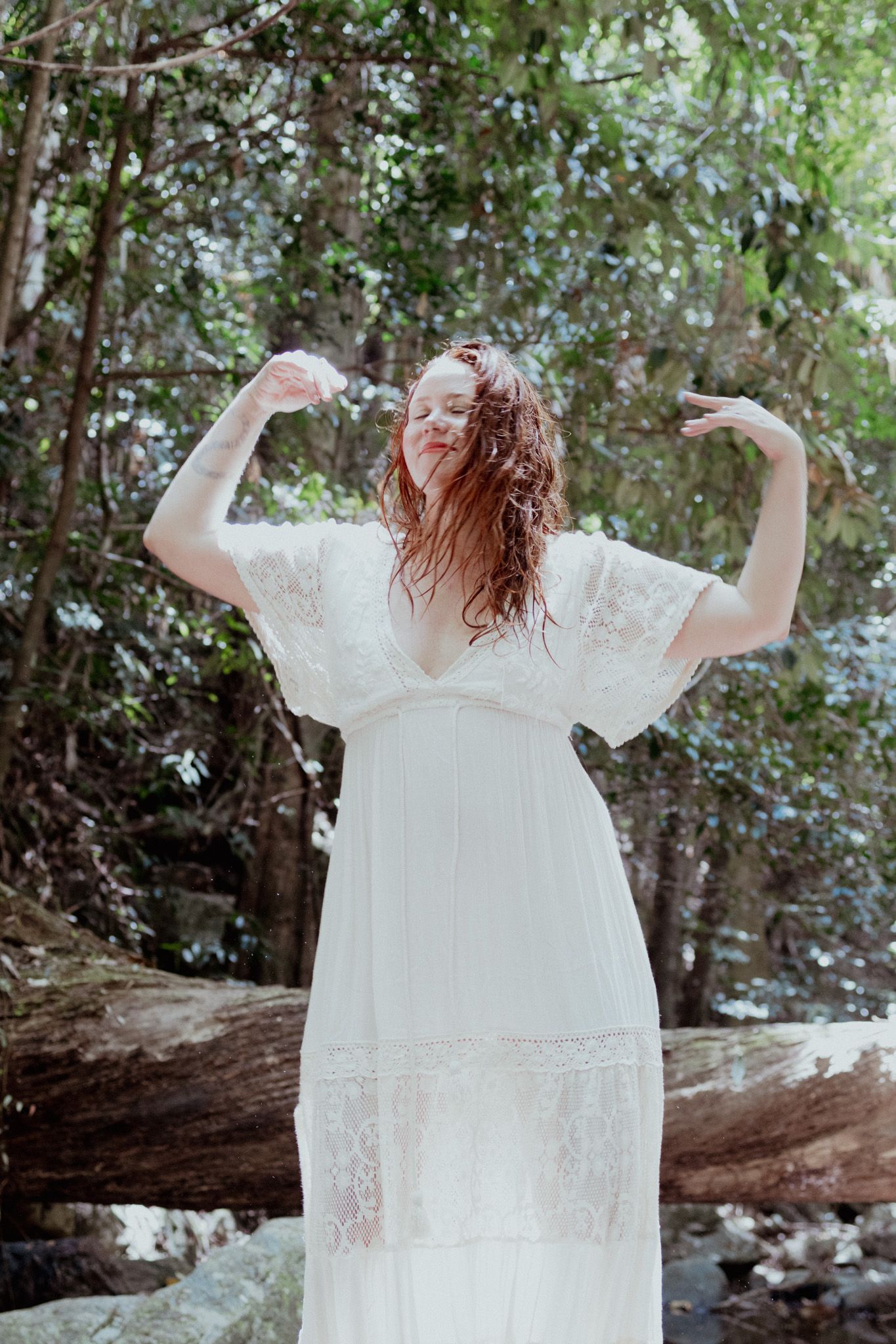 Person in white lace dress poses in natural woodland setting with dramatic lighting.