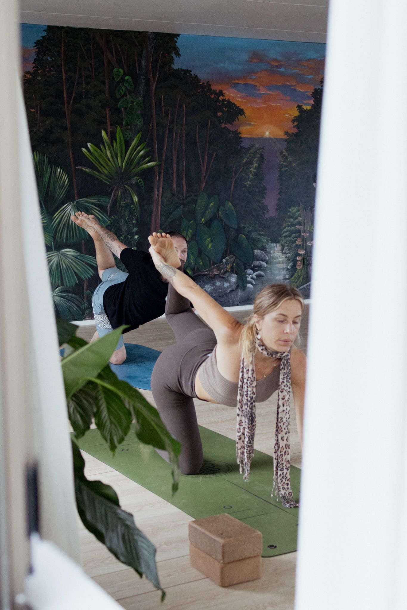 Two people practice yoga on green mats in a bright studio with a tropical jungle mural, a wooden block, and leafy plants in the foreground.