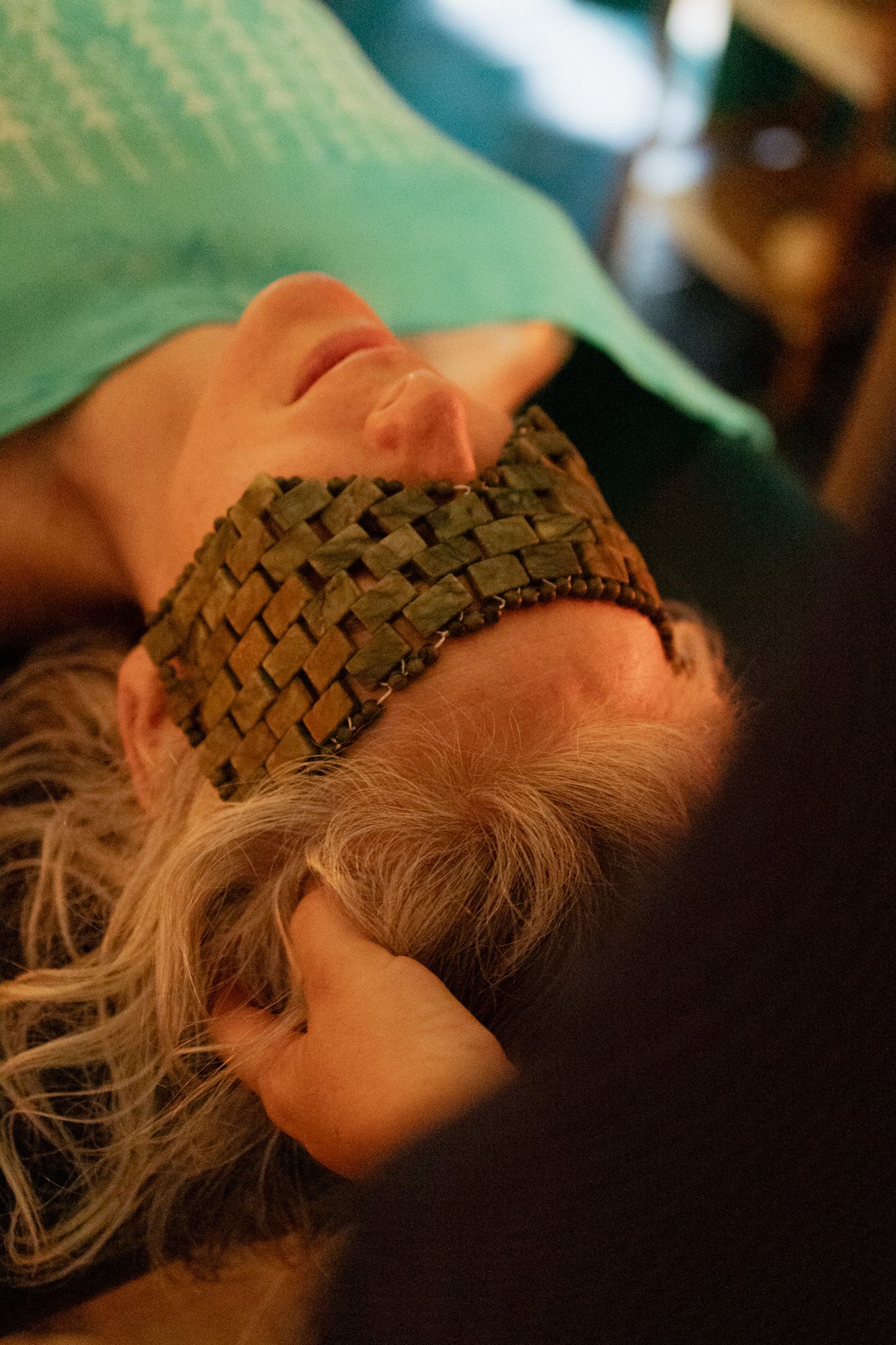 Woman lying down wearing a woven beaded eye mask while a therapist gently holds her head during a relaxing wellness treatment.
