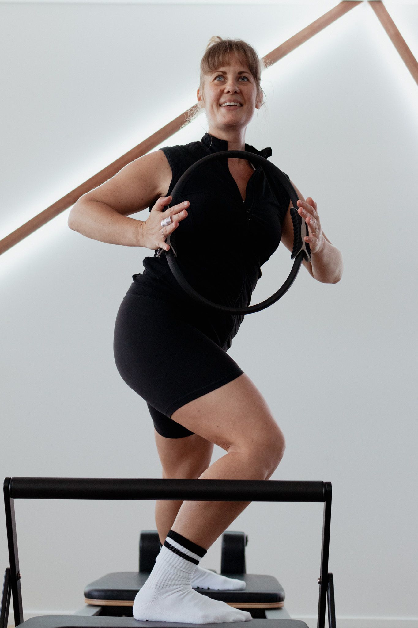 A smiling woman in black athletic wear holds a Pilates ring while standing on a Pilates reformer in a bright studio.