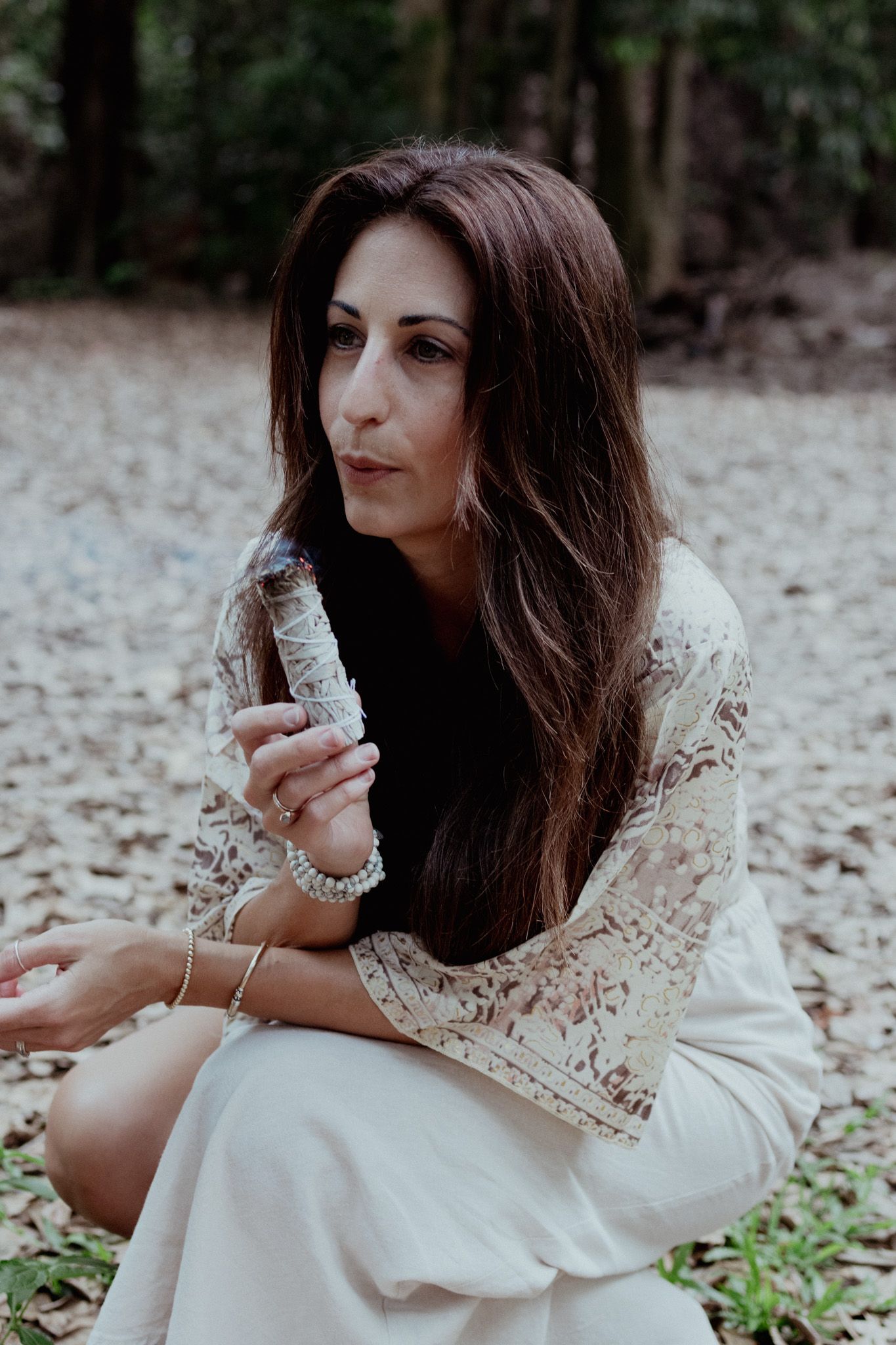 Woman in white lace dress poses elegantly on forest floor.