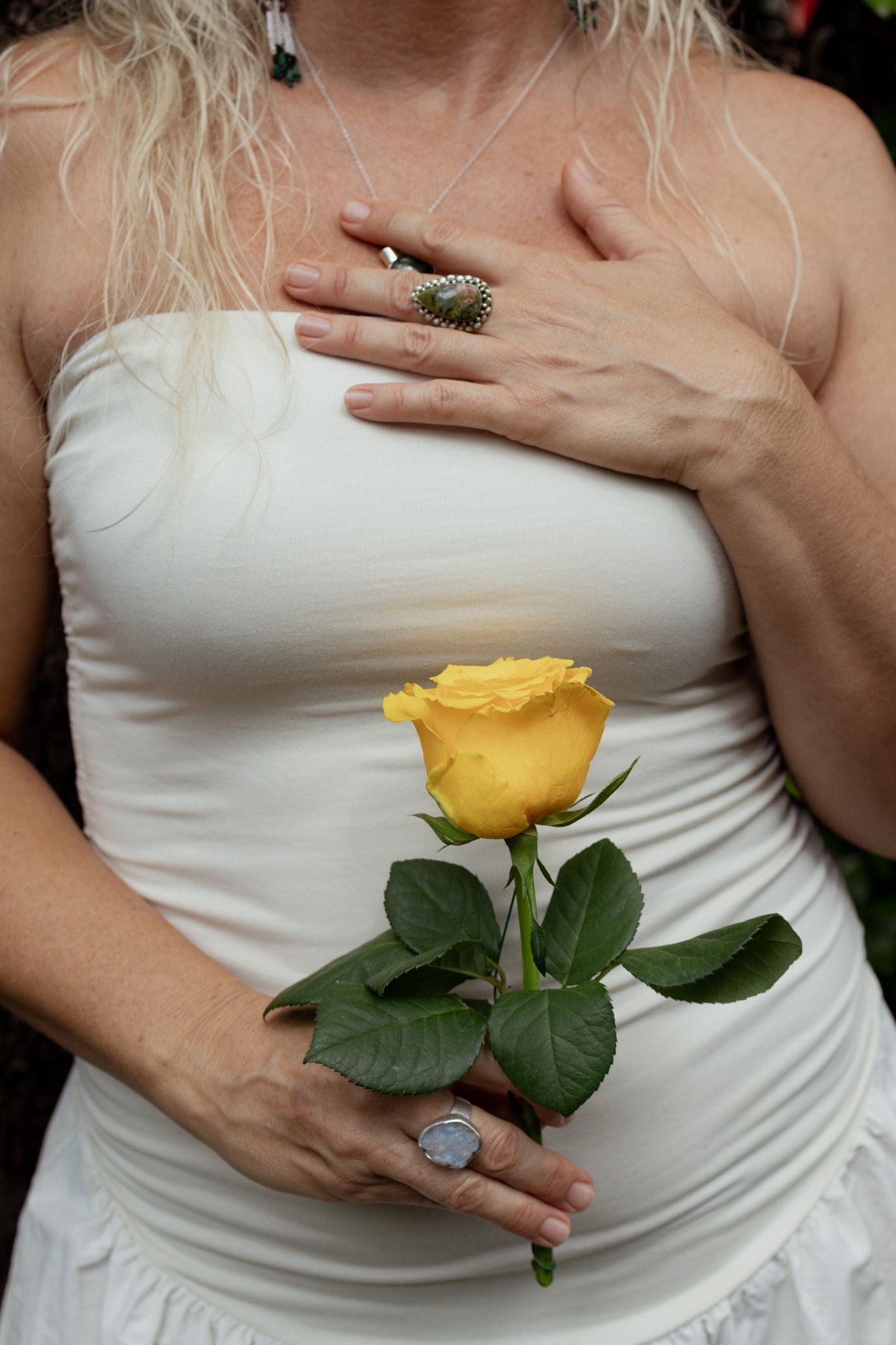 Woman in a white strapless top holding a single yellow rose, one hand resting on her chest, wearing silver rings and a necklace.