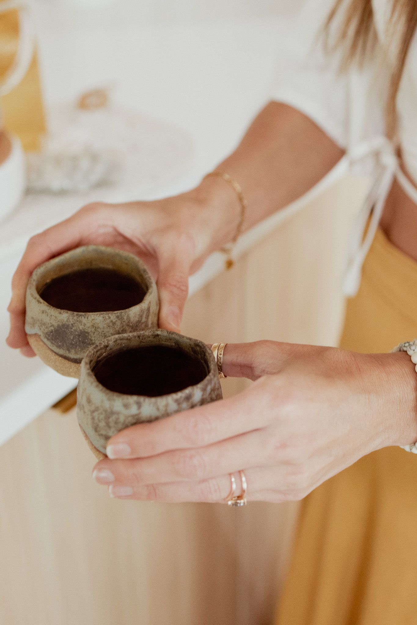 Woman in a mustard skirt and white top holds two rustic handmade ceramic cups filled with dark coffee.