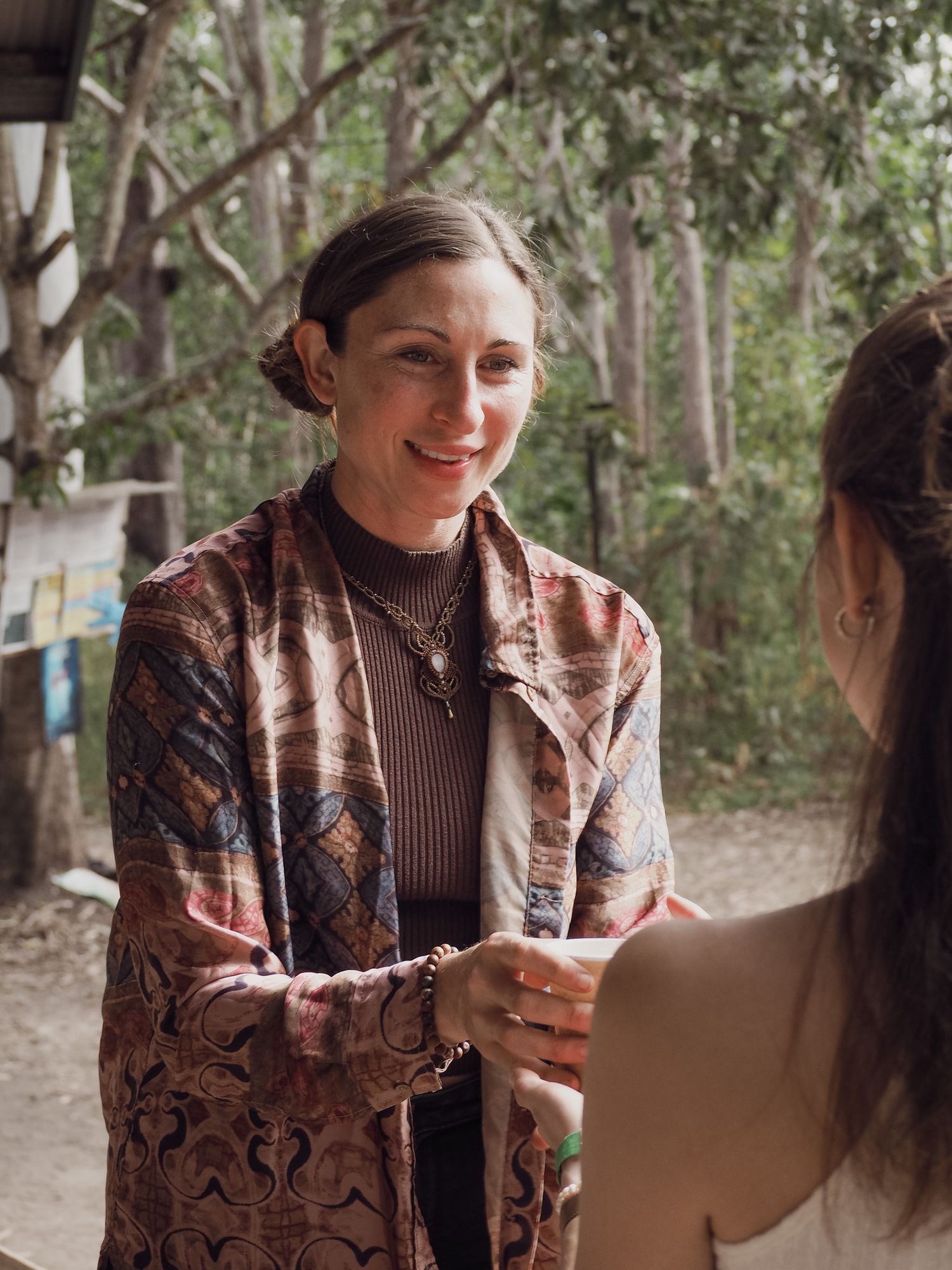 Person in patterned jacket having conversation at outdoor gathering with eucalyptus trees behind.