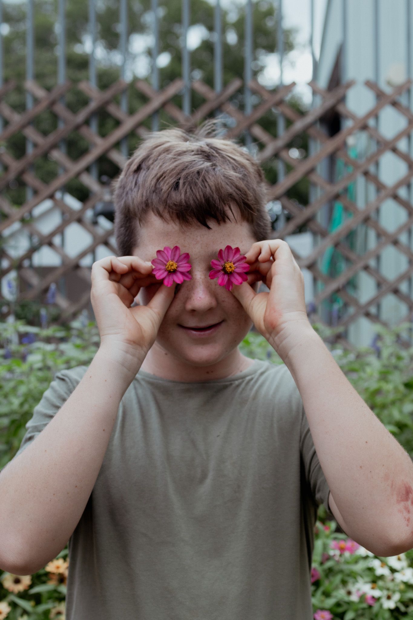 Child playfully holding pink flowers over their eyes in a garden setting.