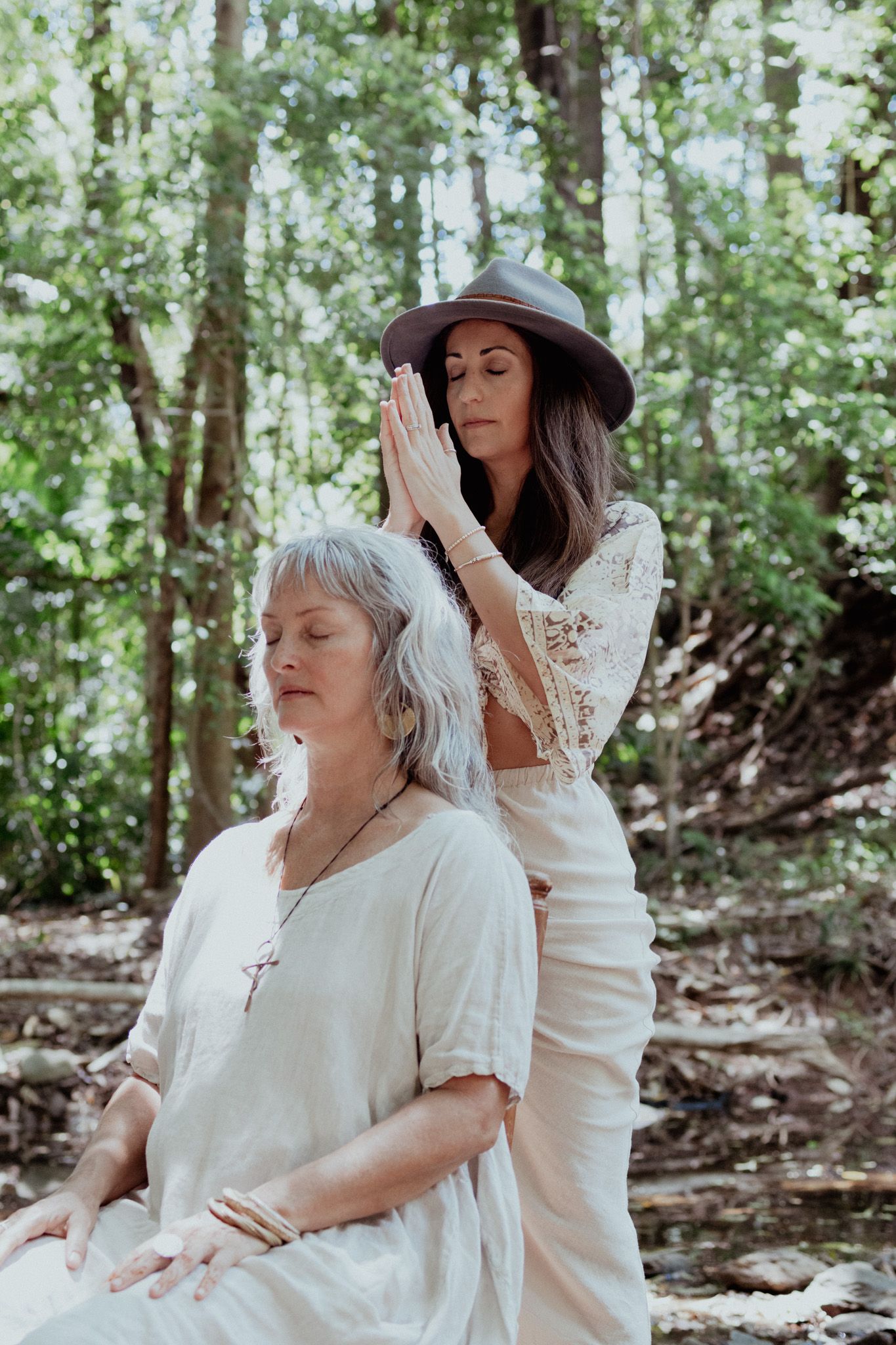 Two people in white dresses share an intimate moment in a sunlit forest setting.