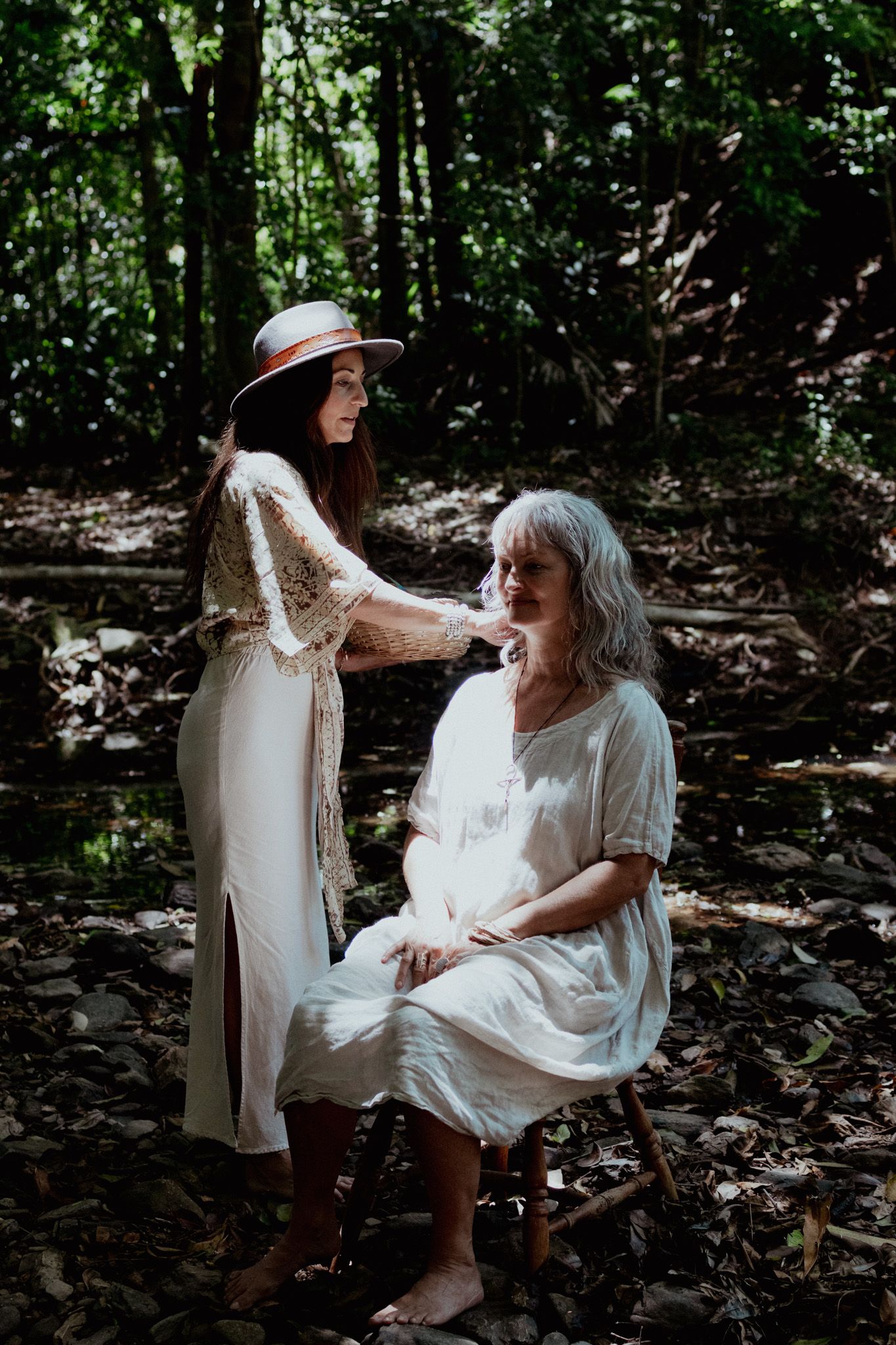 Two people share a tender moment in white dresses against a forest backdrop.
