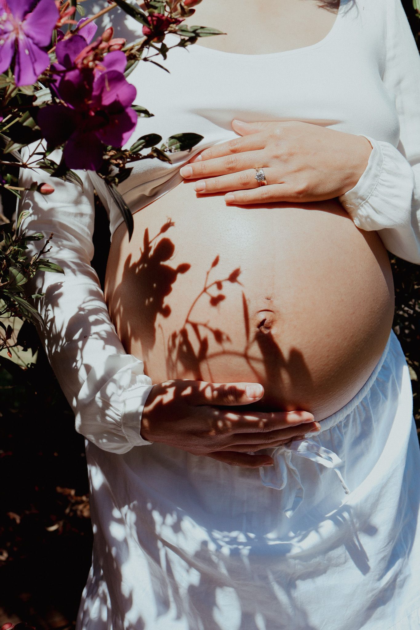 Maternity photos showing a pregnant belly with dappled sunlight and purple flowers in the background.