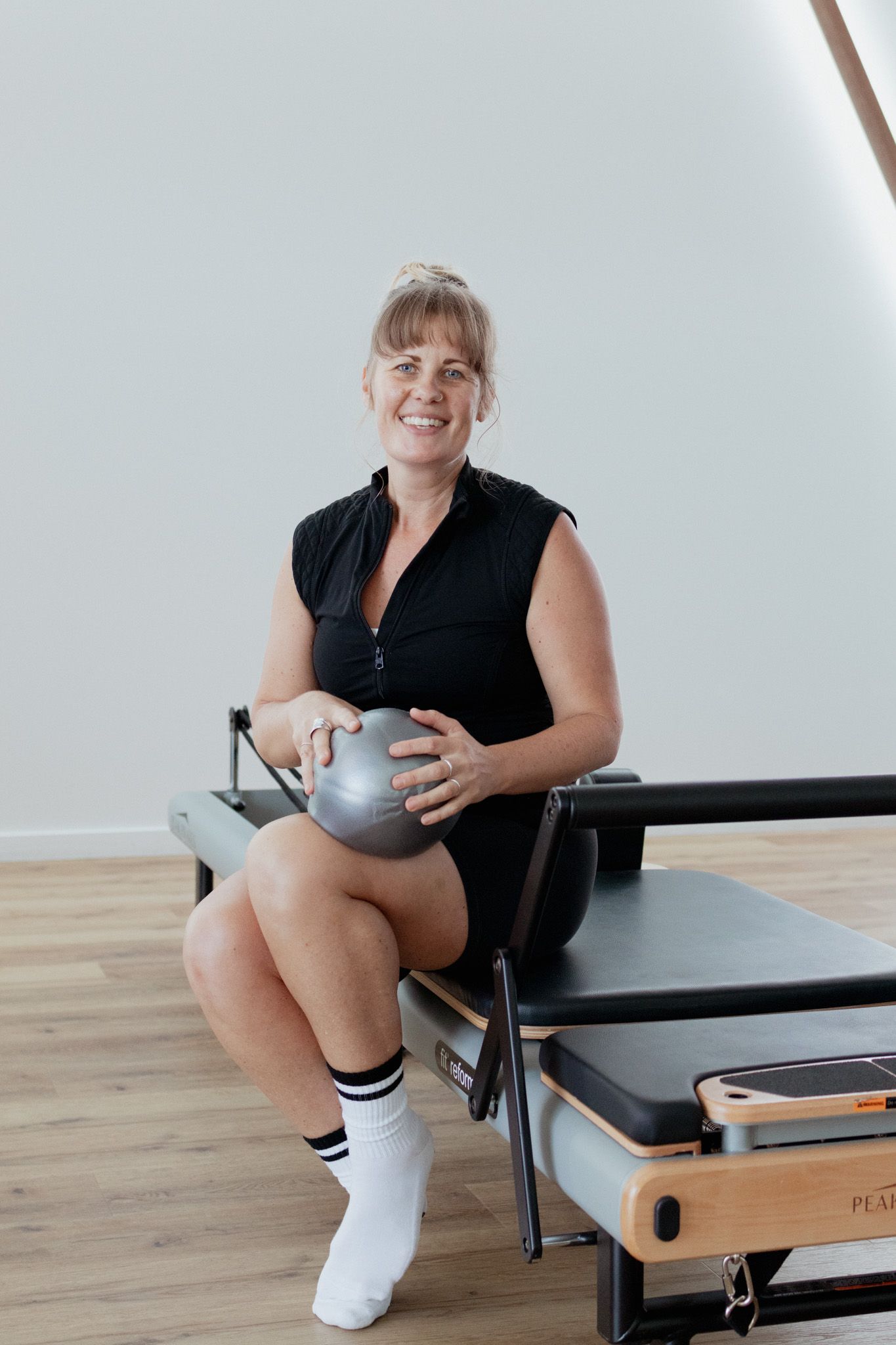 Smiling woman in a black sleeveless top and white socks sits on a Pilates reformer holding a small gray exercise ball in a bright studio.