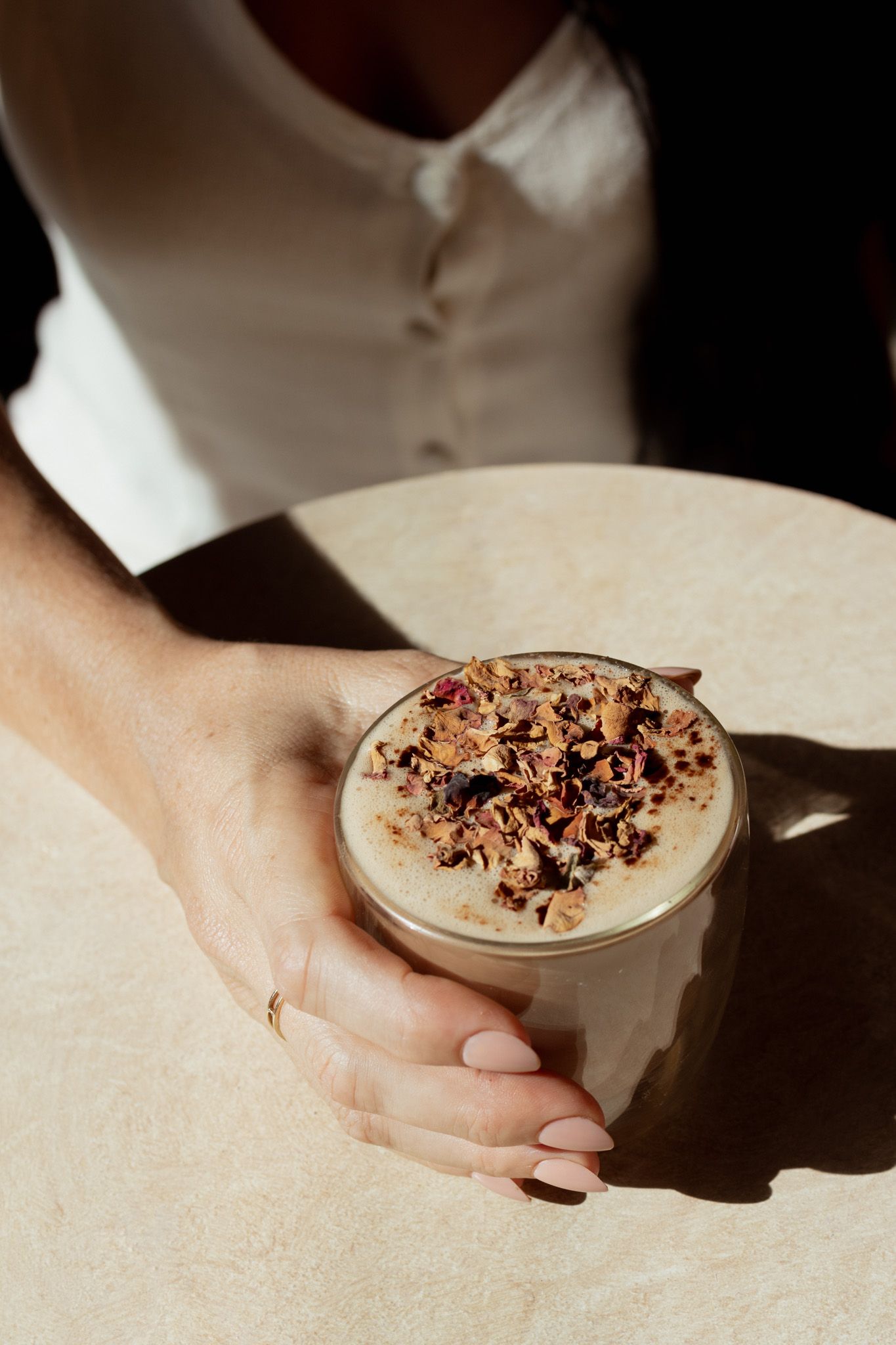 Woman in a white blouse holding a glass of creamy latte topped with dried rose petals and nuts on a sunlit table.