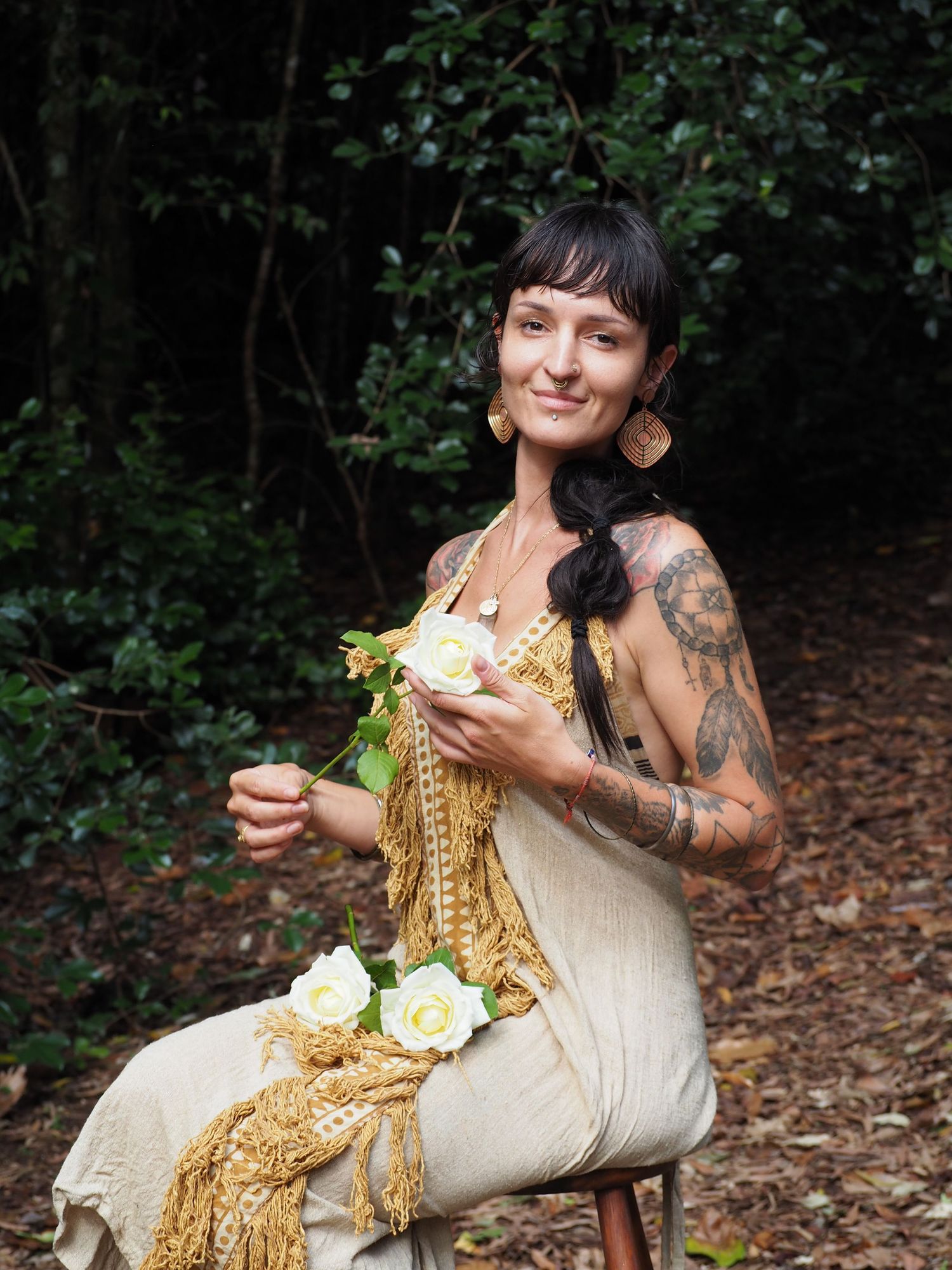 A person in a white dress with gold accents sits outdoors holding flowers.