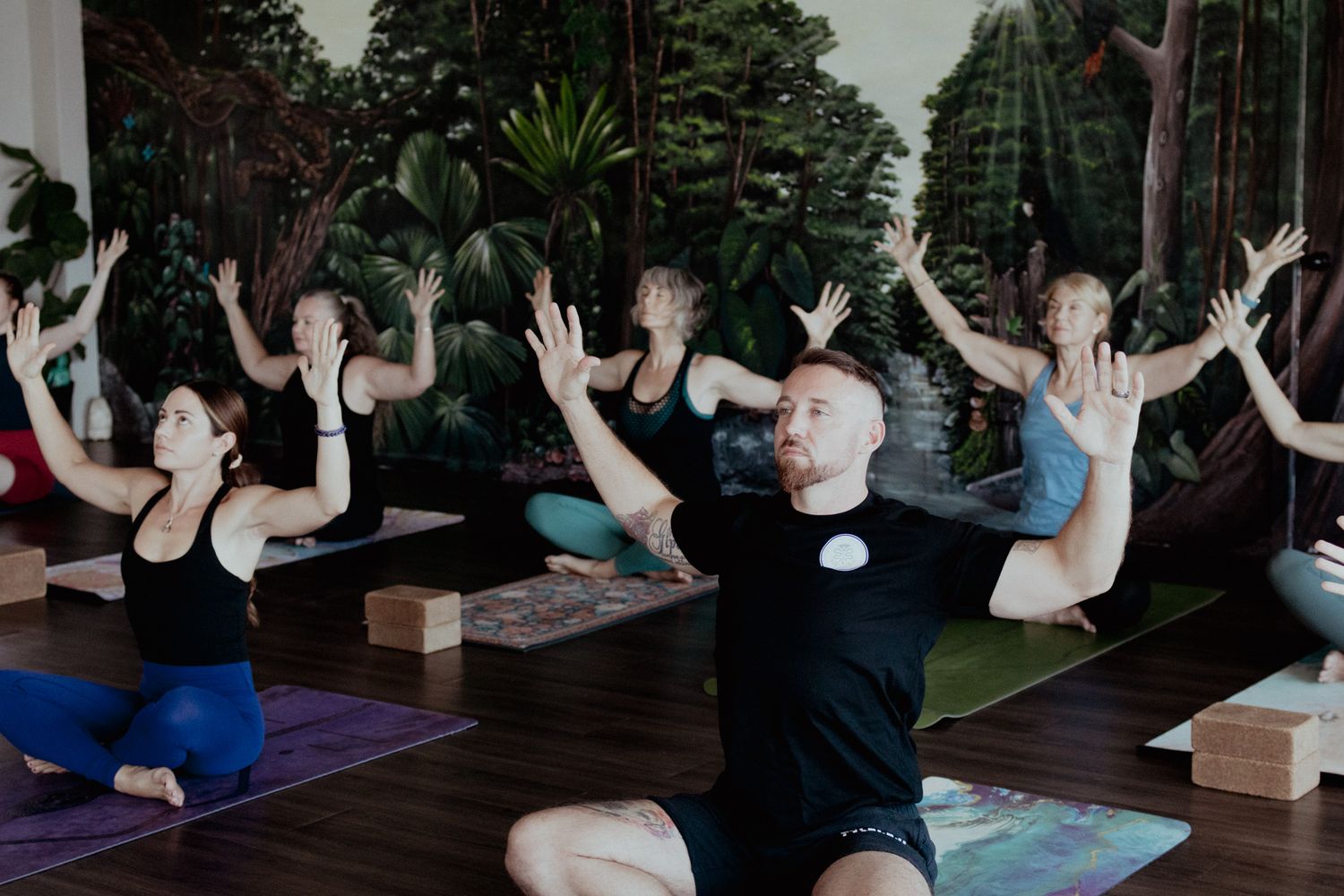 Group yoga class with participants raising arms in synchronization on yoga mats.