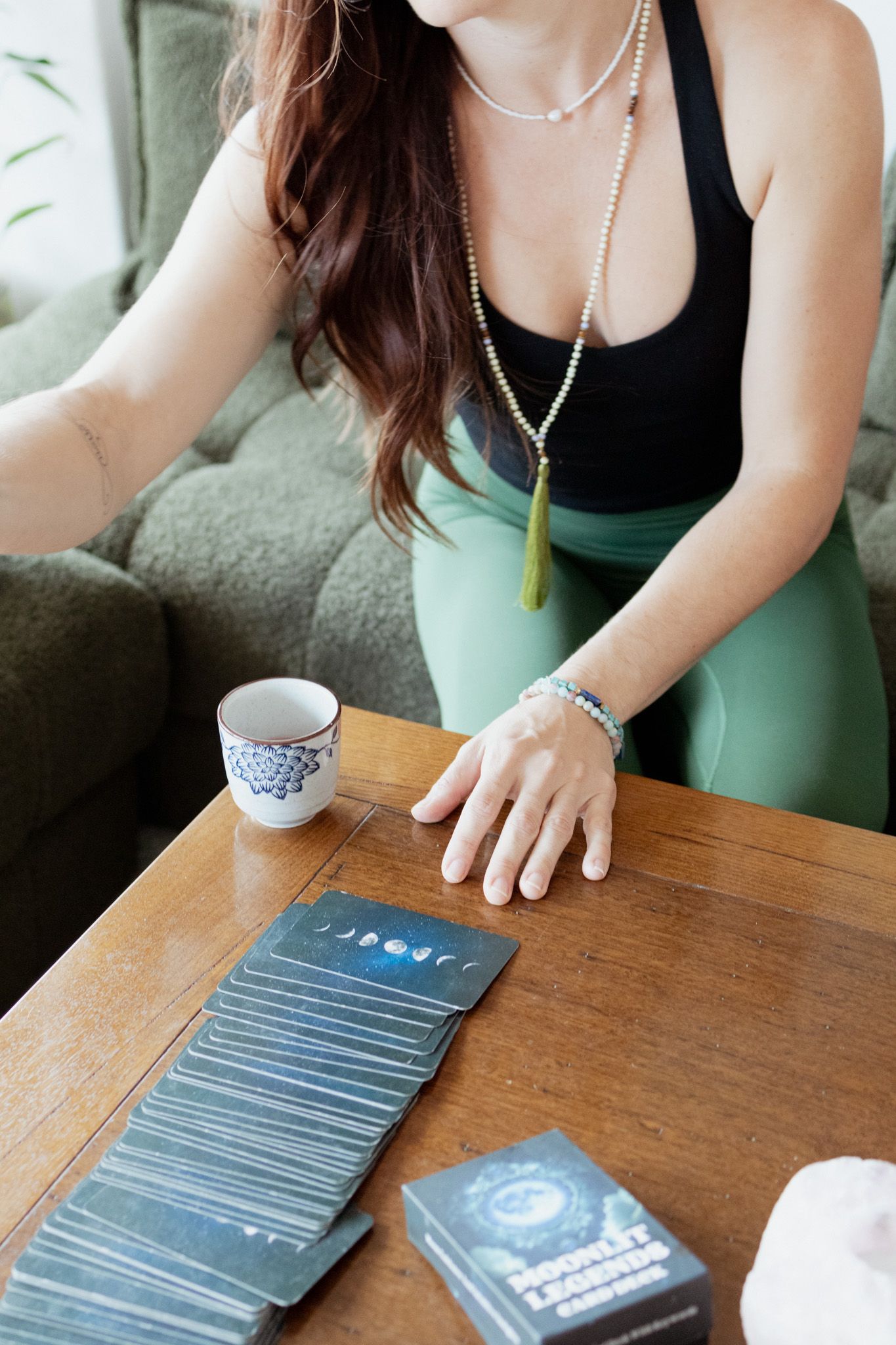 Woman in green leggings and a black tank top lays out a fanned row of moon-themed tarot cards on a wooden table beside a floral teacup.