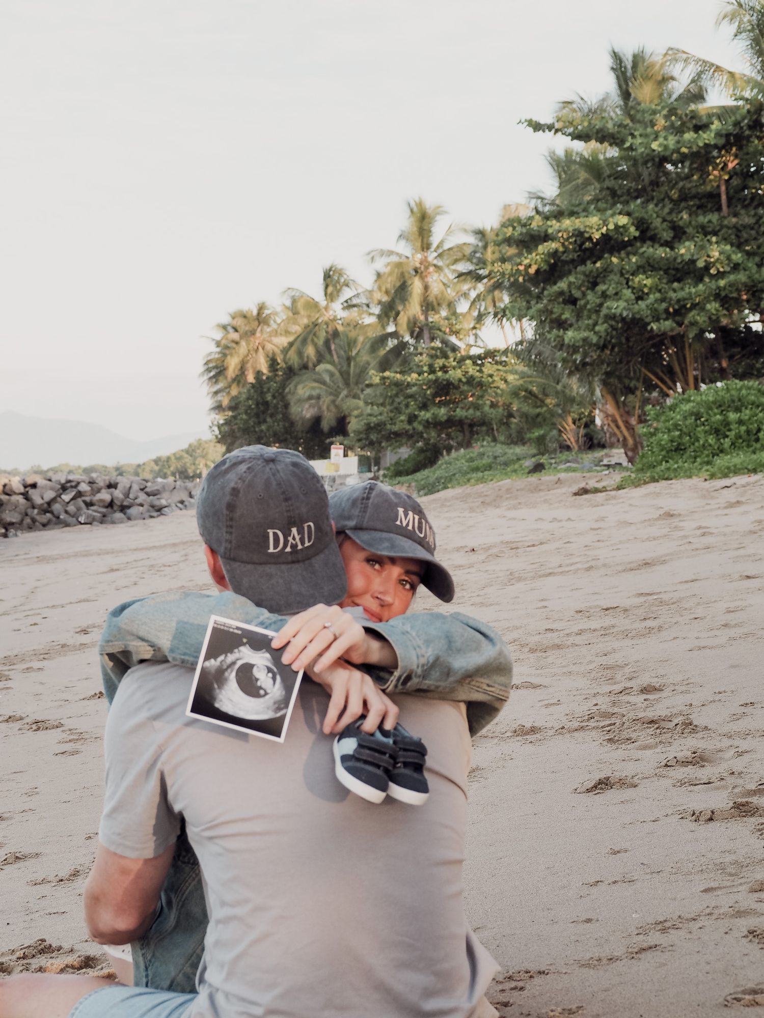 Couple embracing on beach wearing matching DAD and MUM caps with ultrasound photo.