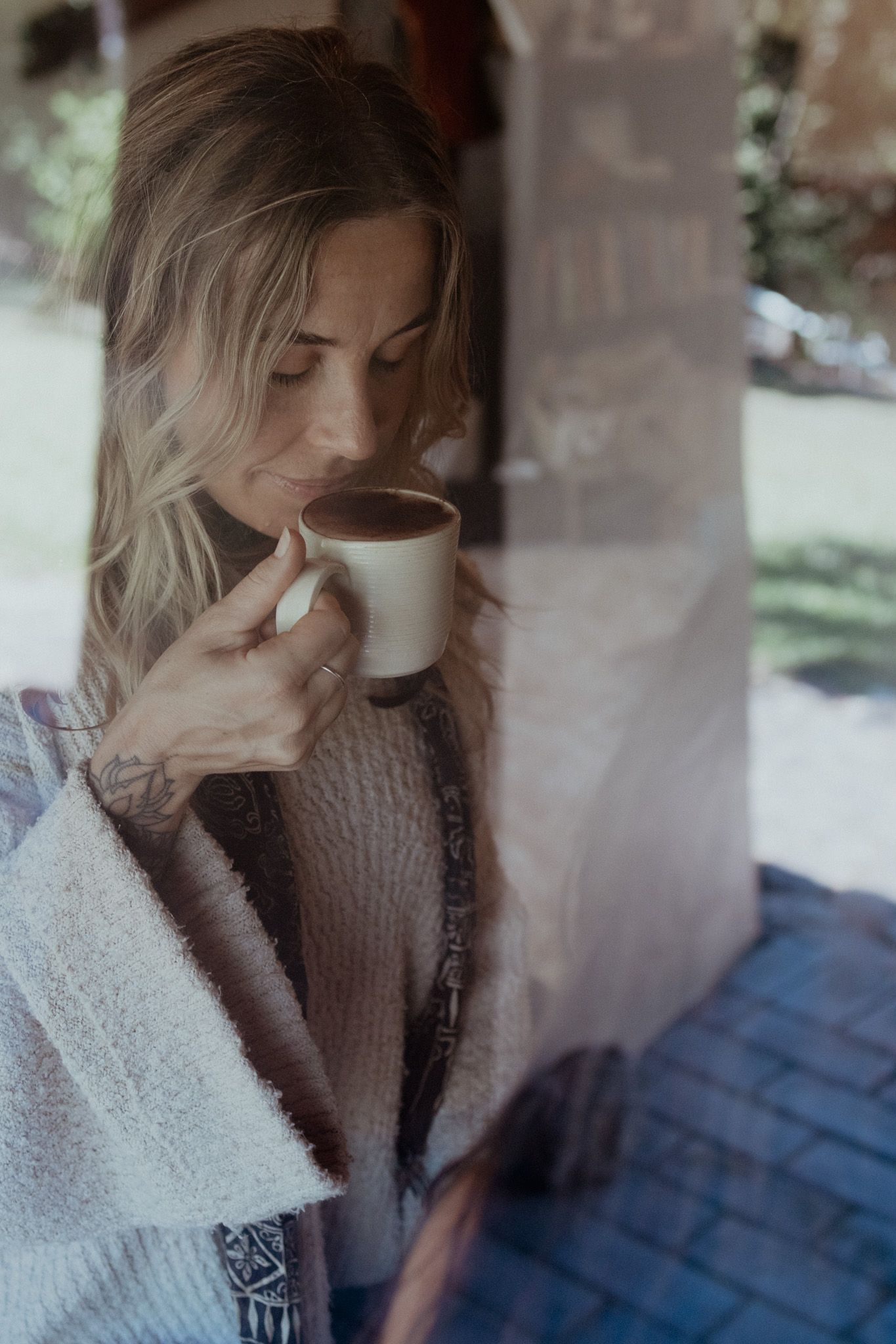 Person in cozy sweater drinking from a mug by a window.