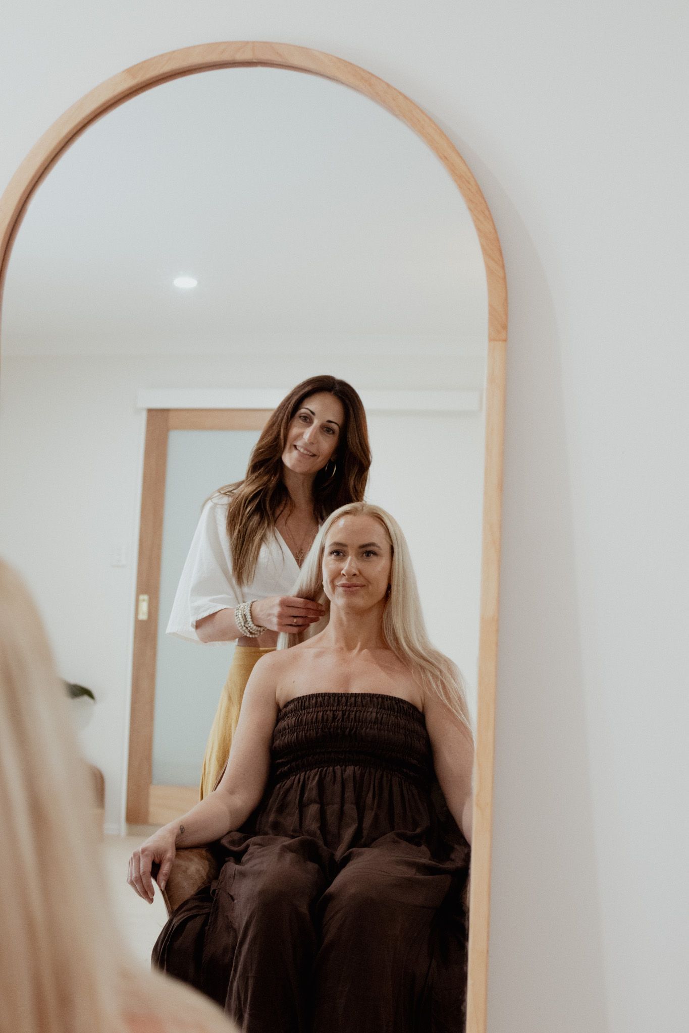 Mirror reflection of a smiling hairstylist styling the long blonde hair of a seated woman in a brown dress inside a bright salon.