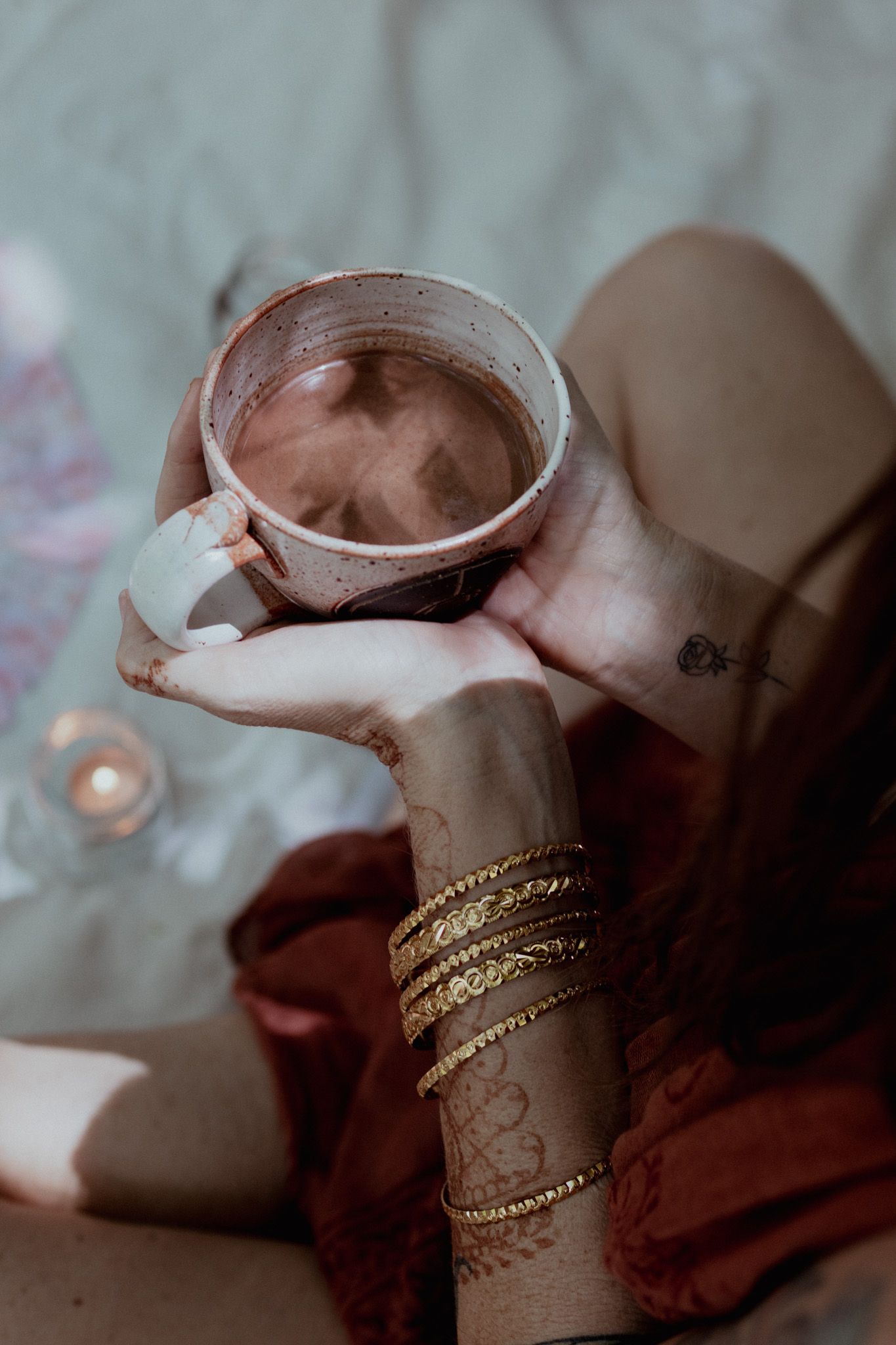 Artistic photo of hands wearing gold bracelets holding ceramic cup with moody lighting.