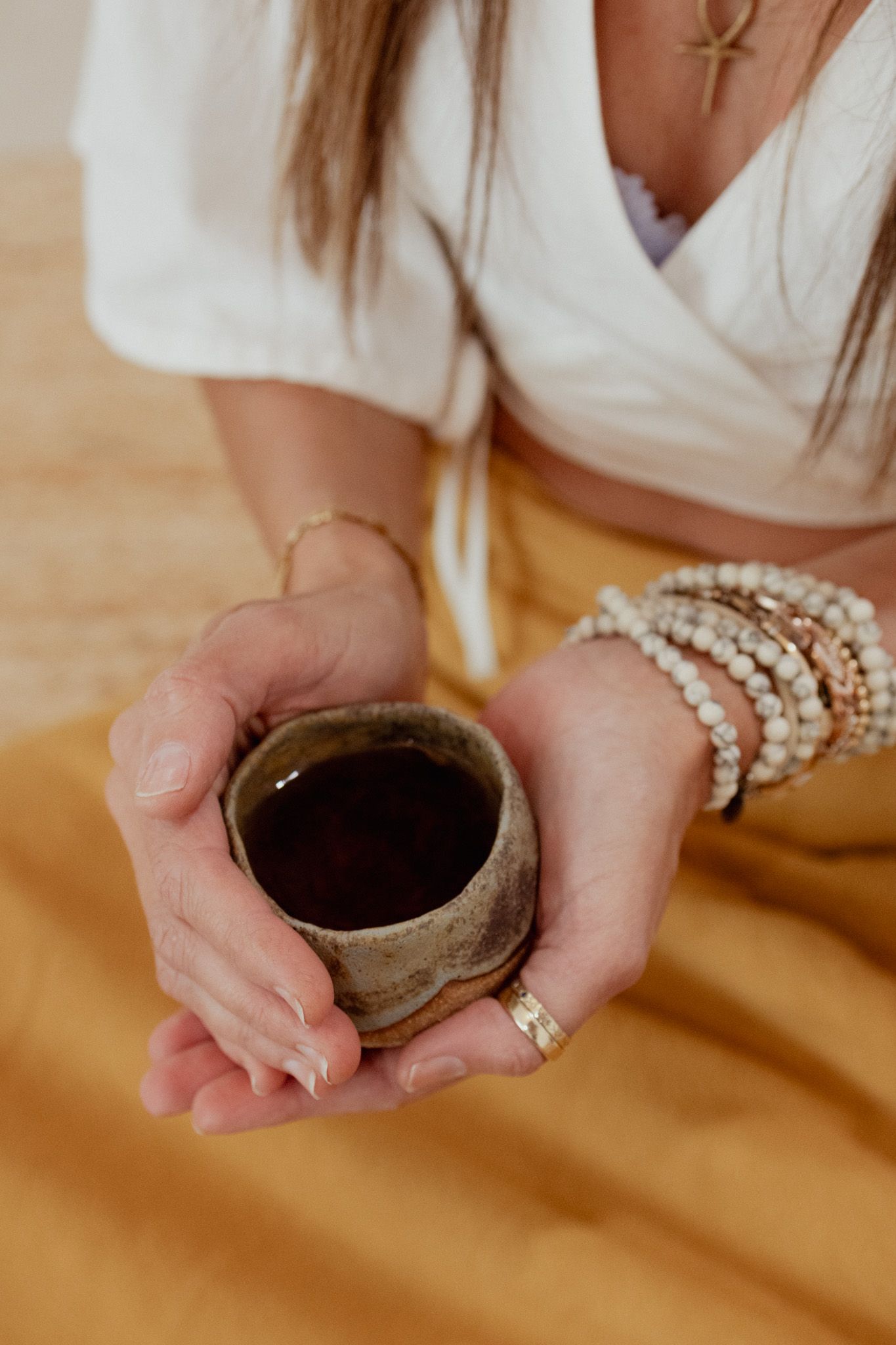 Woman in a white wrap top holds a rustic ceramic cup of dark tea in cupped hands, wearing beaded bracelets and a gold ring.