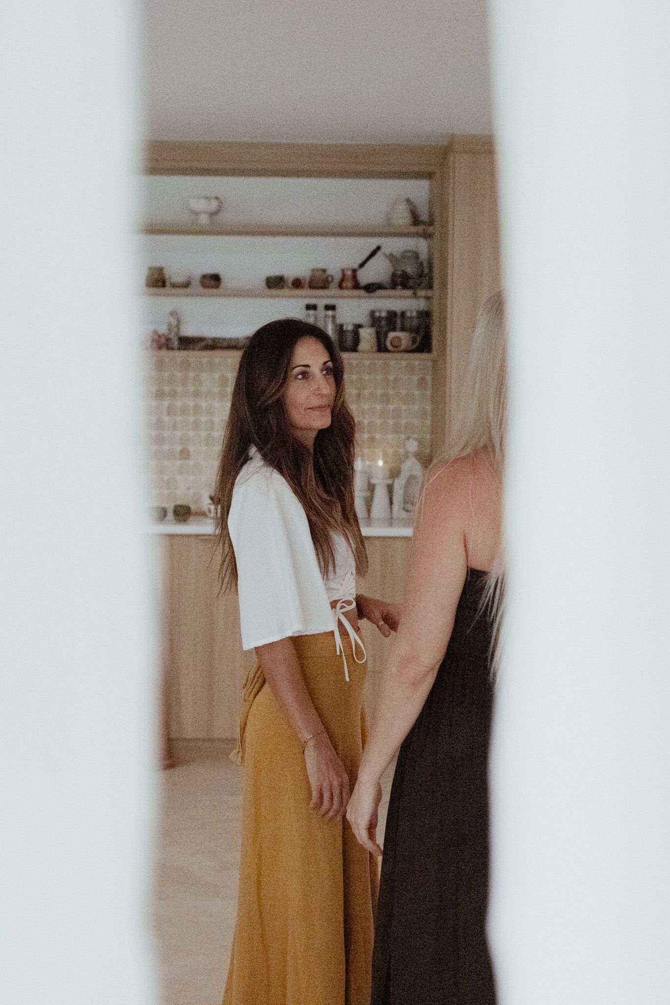 Two women talk in a bright, modern kitchen with wooden shelves, one wearing a white top and mustard skirt, seen through a narrow gap.