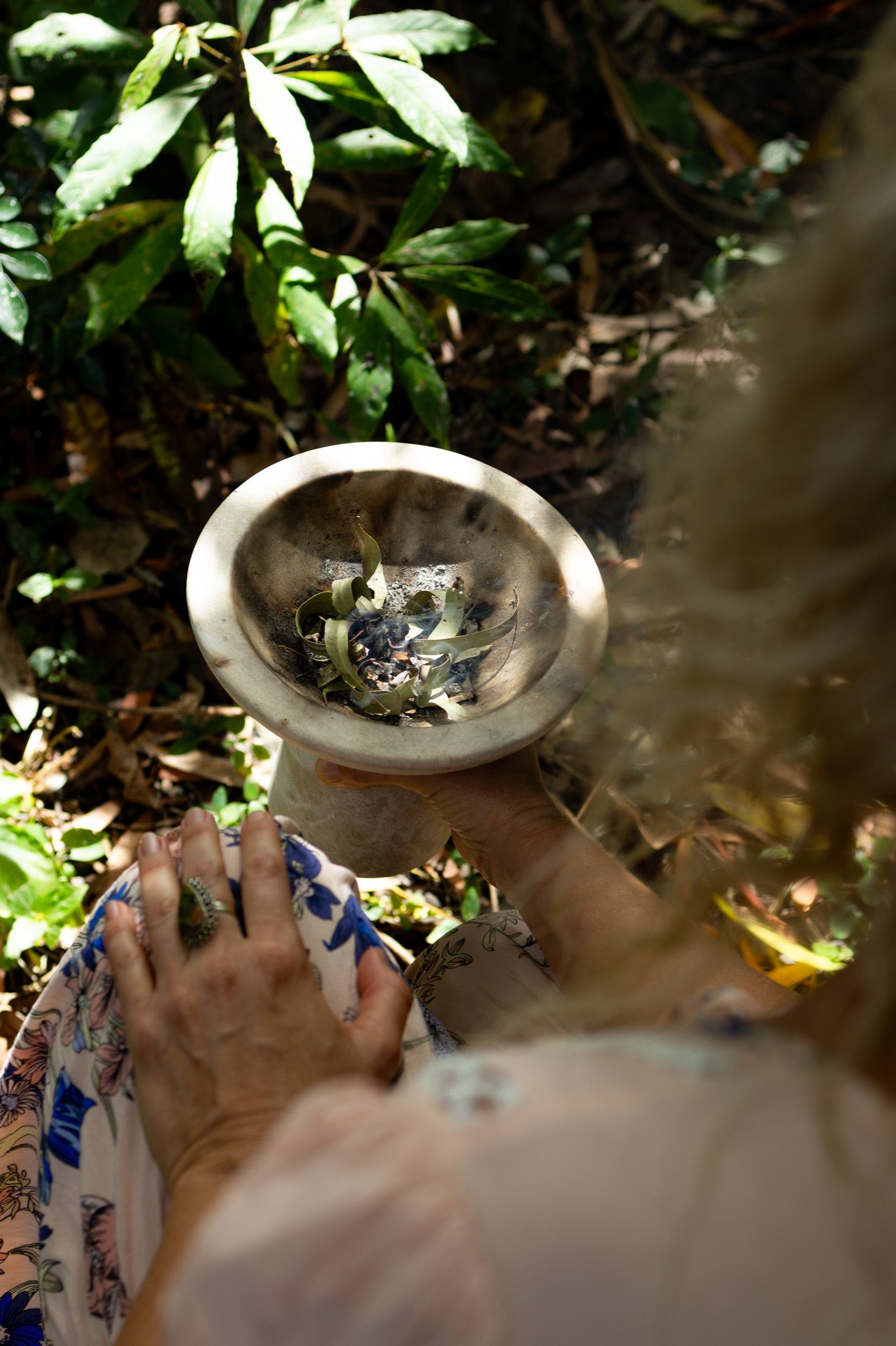 Person in a floral dress holds a stone bowl of burning herbs and leaves during an outdoor smudging ritual surrounded by lush green foliage.